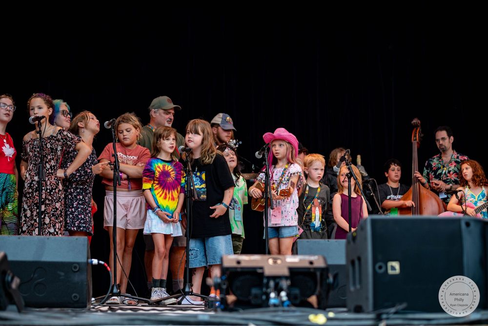 Children and adults perform together on an outdoor stage at a music festival, with microphones and instruments visible.