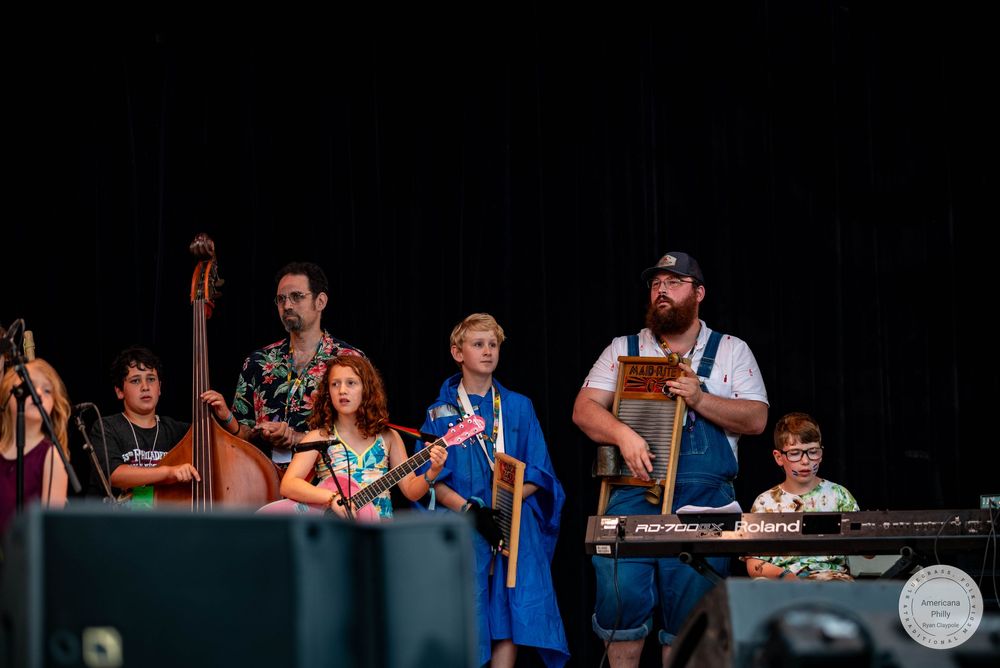 A colorful group of musicians performing on stage outdoors, holding various instruments including a bass and ukulele.