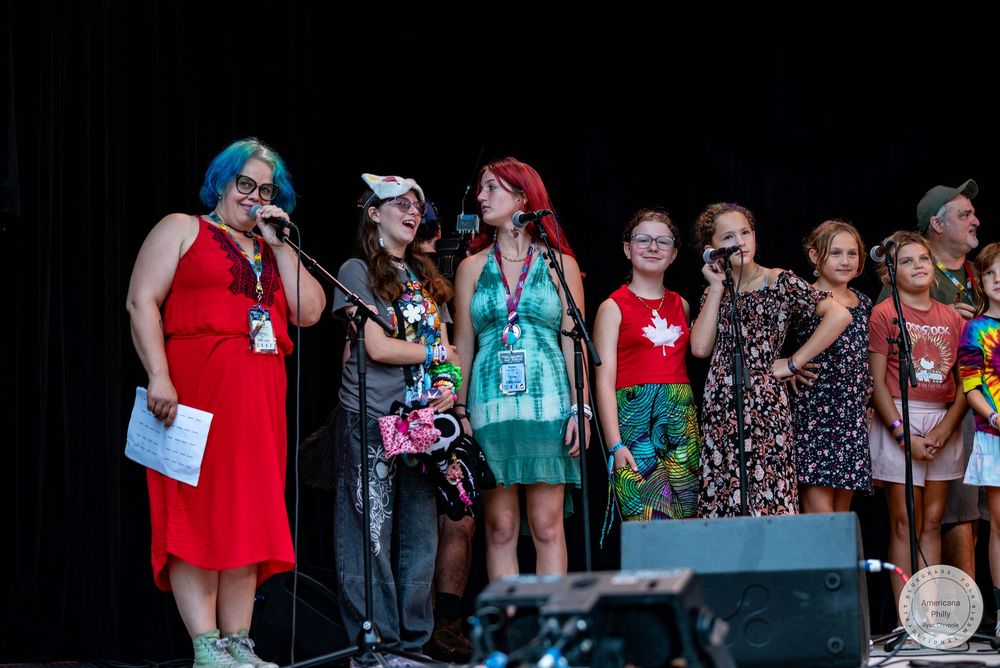Group of women singing on outdoor stage, some with colorful hair, wearing festive attire at a summer event.
