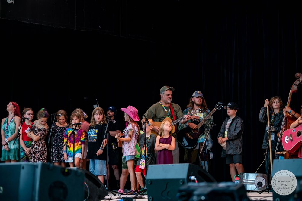 Children and adults perform together on an outdoor festival stage with guitars and instruments under bright lights.