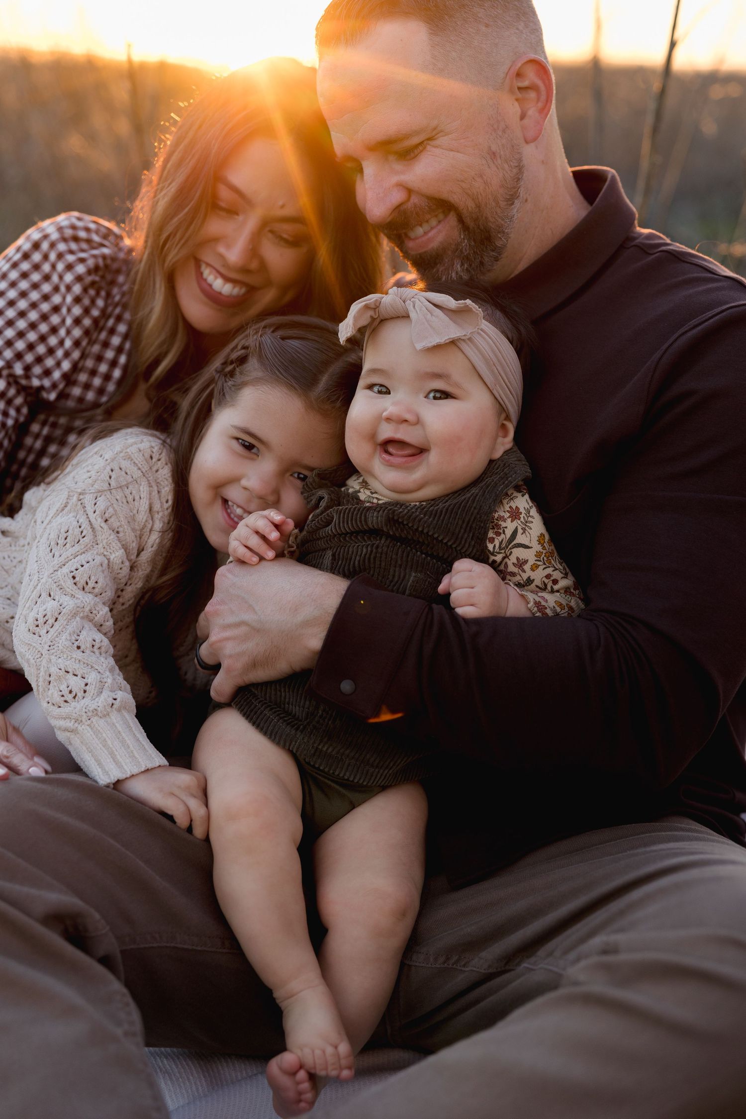 Happy family of four sharing a warm moment at golden hour sunset, with parents smiling at their two young daughters outdoors.