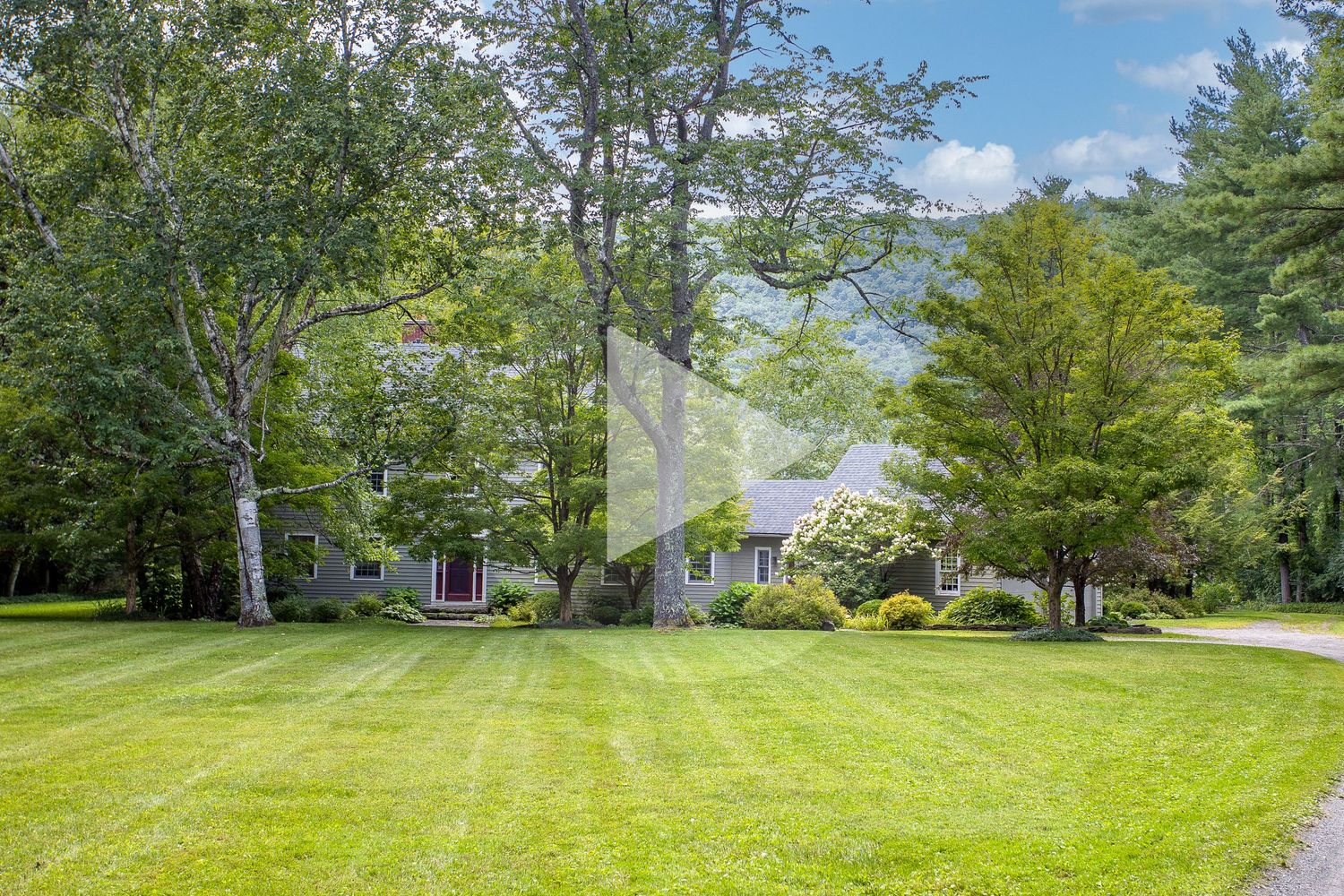 Cape Cod style home with manicured lawn surrounded by tall trees and mountains.