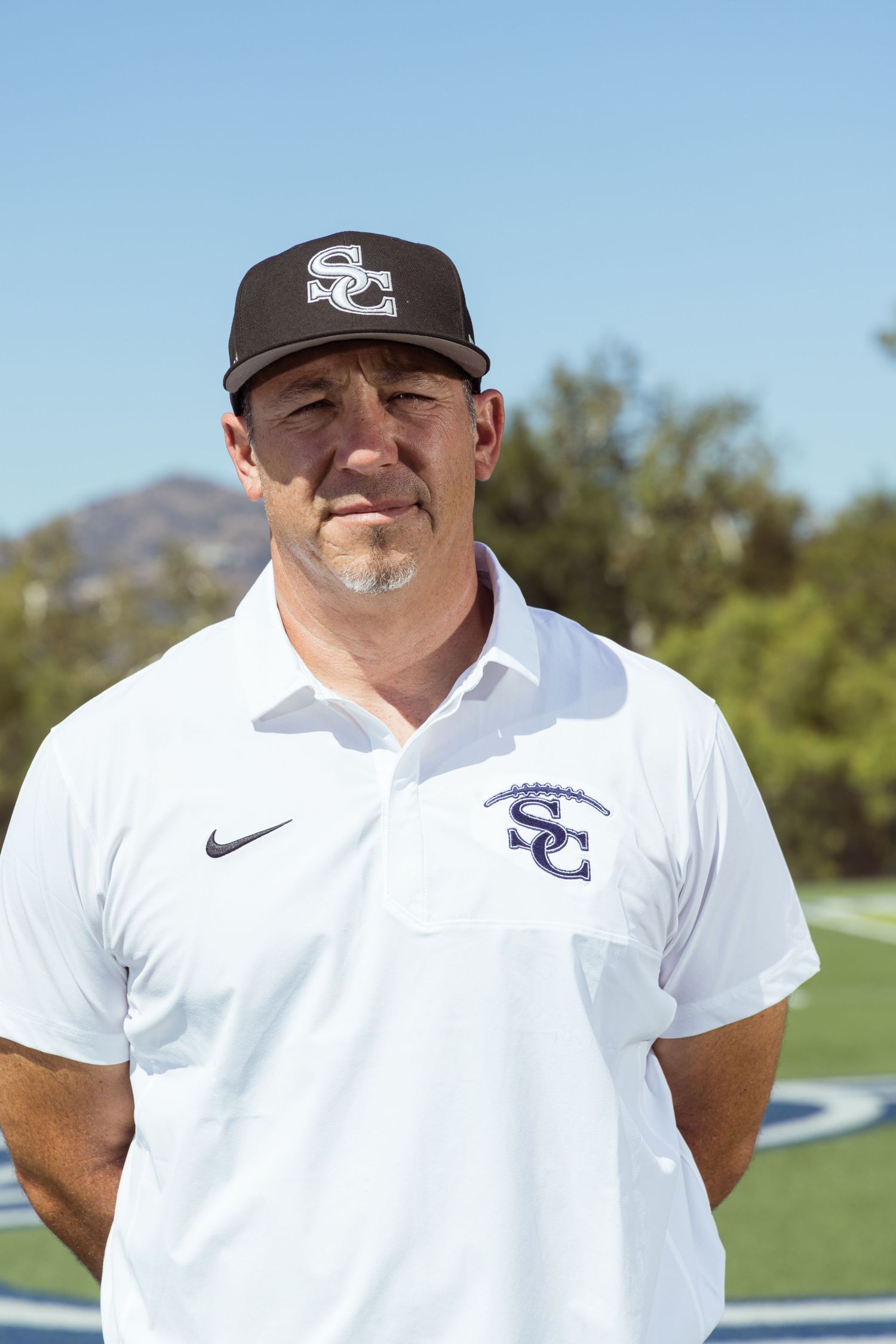 Coach wearing SC baseball cap and white Nike polo shirt stands on sports field with mountains in background.
