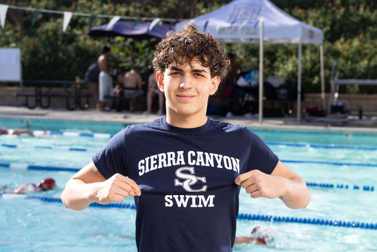 Young swimmer proudly displaying Sierra Canyon Swim team shirt at outdoor pool during swim meet.