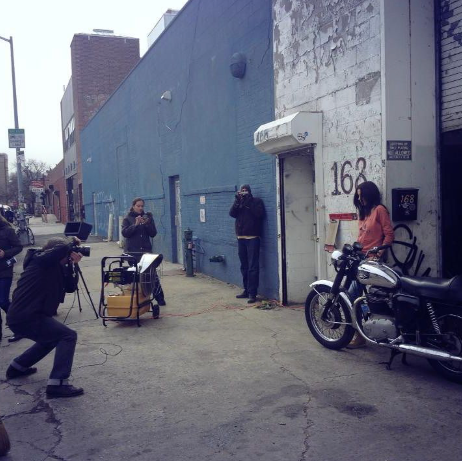 Film crew shooting on urban sidewalk beside blue building with motorcycle and number 163 visible.