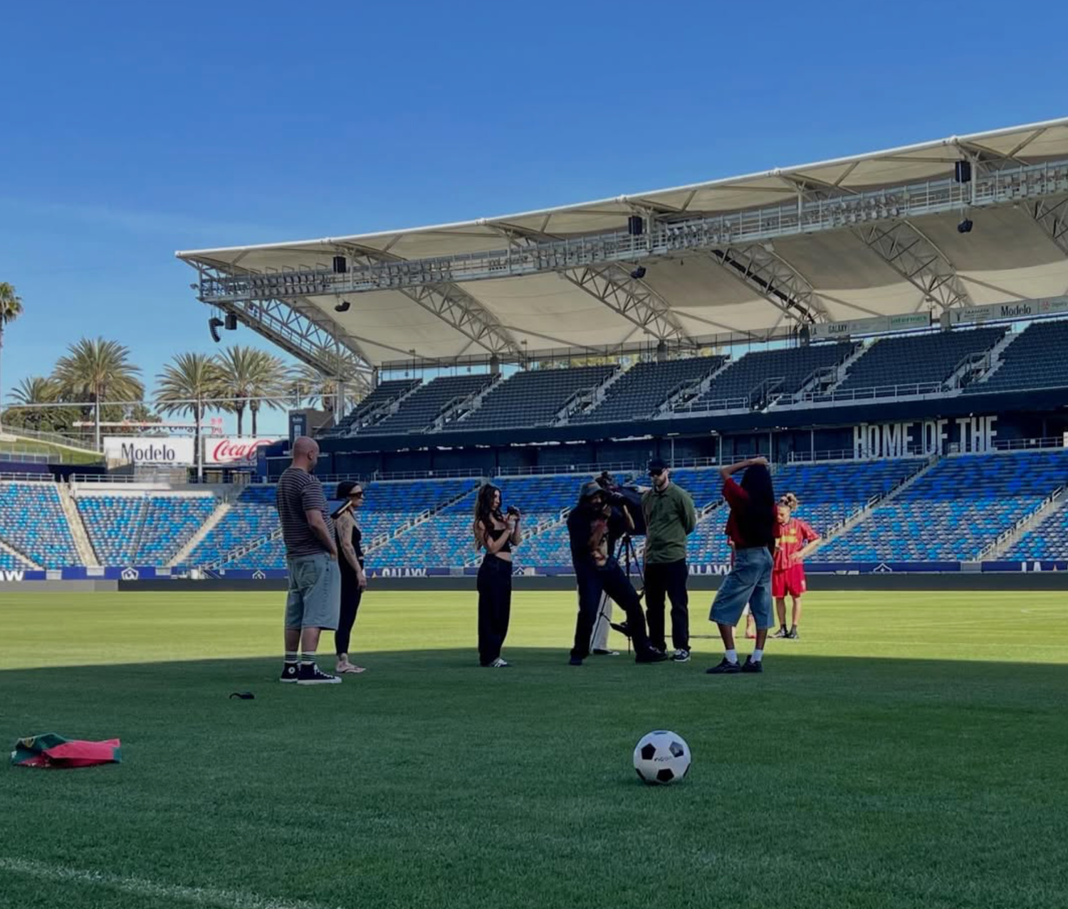 Group of people standing on a soccer field inside a stadium with blue seats and a soccer ball in the foreground.