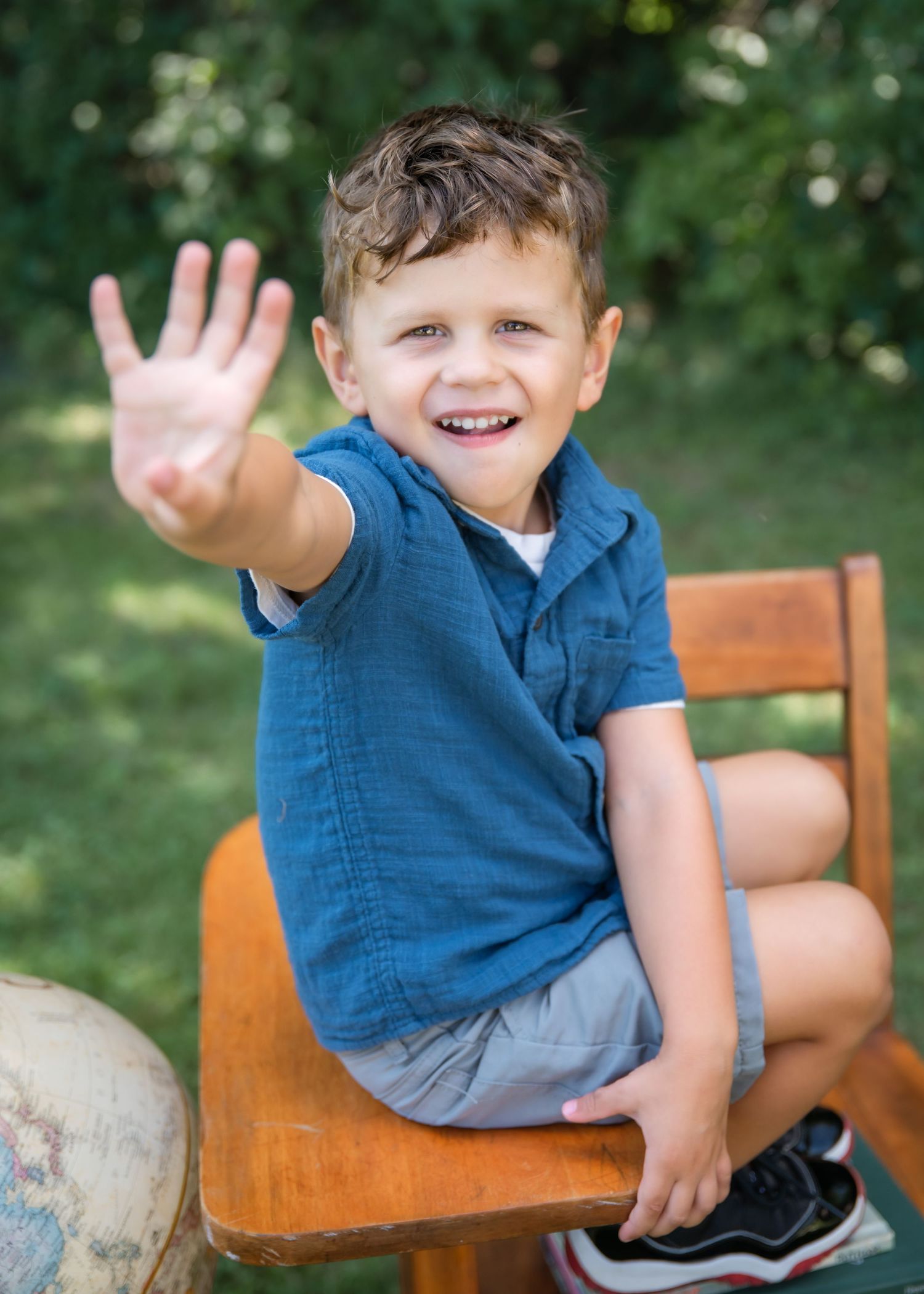 A joyful young boy jumps in the air outdoors, wearing a blue shirt and khaki shorts, with a globe and chair behind him.