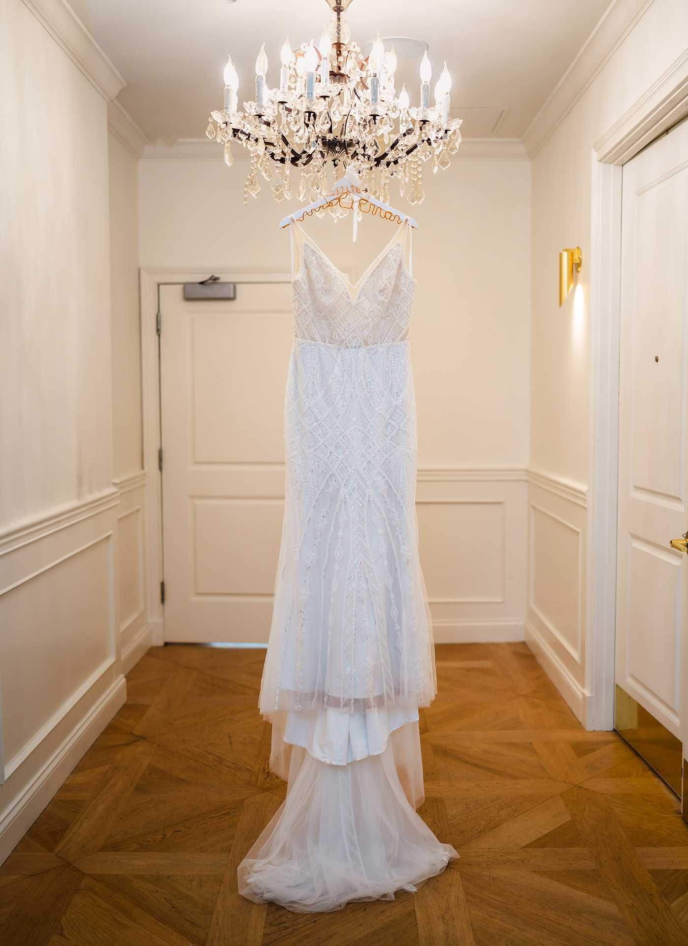 Elegant beaded white wedding gown hanging from a crystal chandelier in the view venue