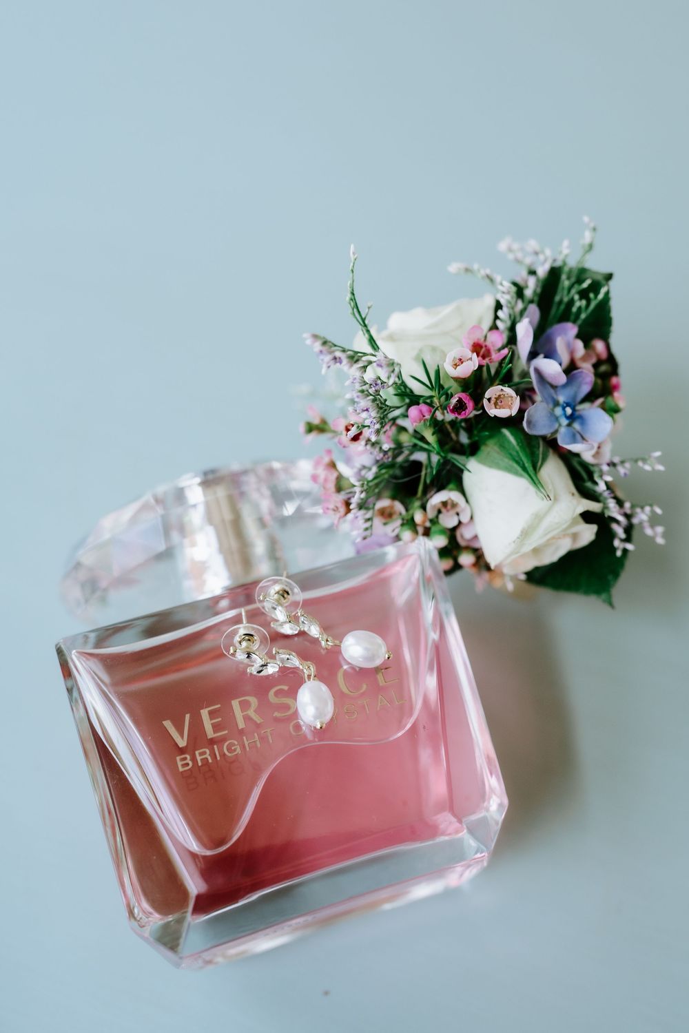 Versace Bright Crystal perfume bottle with pearl earrings and a small floral bouquet on a light blue surface.