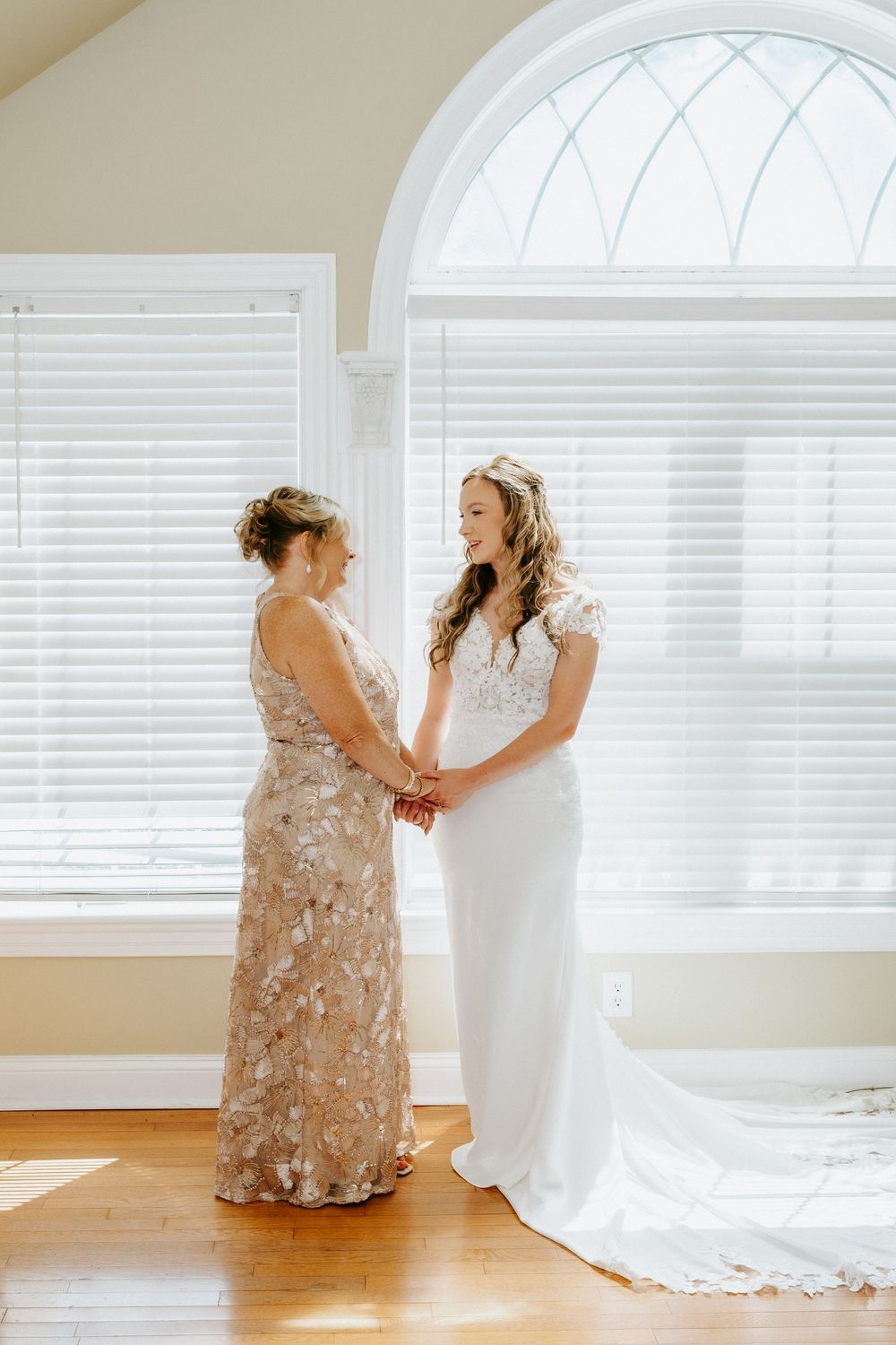 Bride in white lace gown holds hands with woman in gold floral dress near arched window on wedding day.