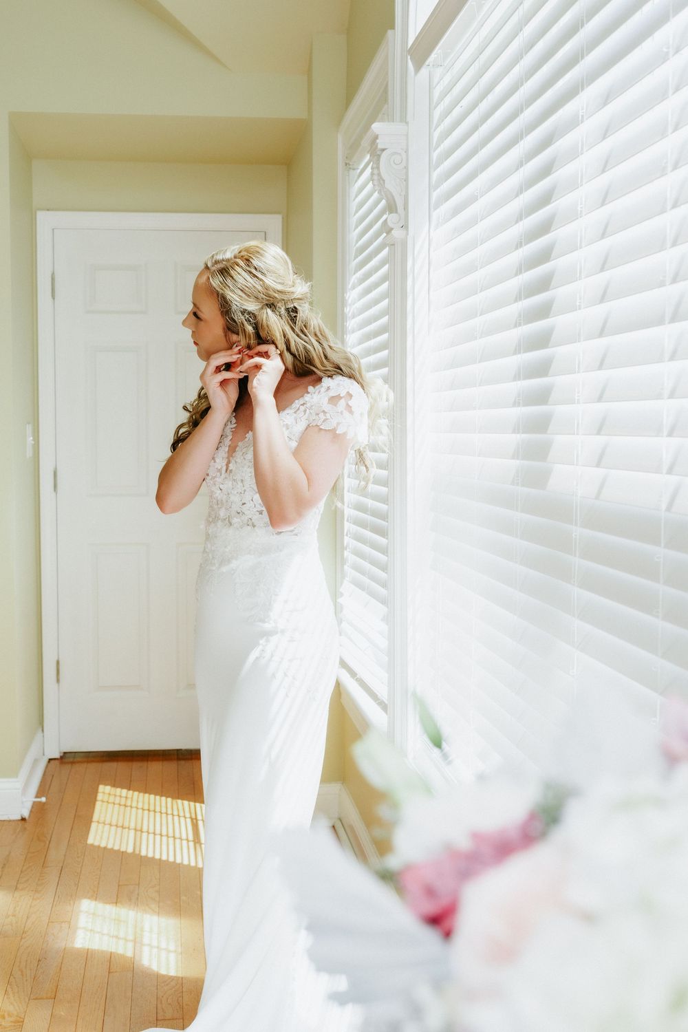 Bride in white lace wedding gown putting on earrings by a sunlit window with a pink bouquet in the foreground.
