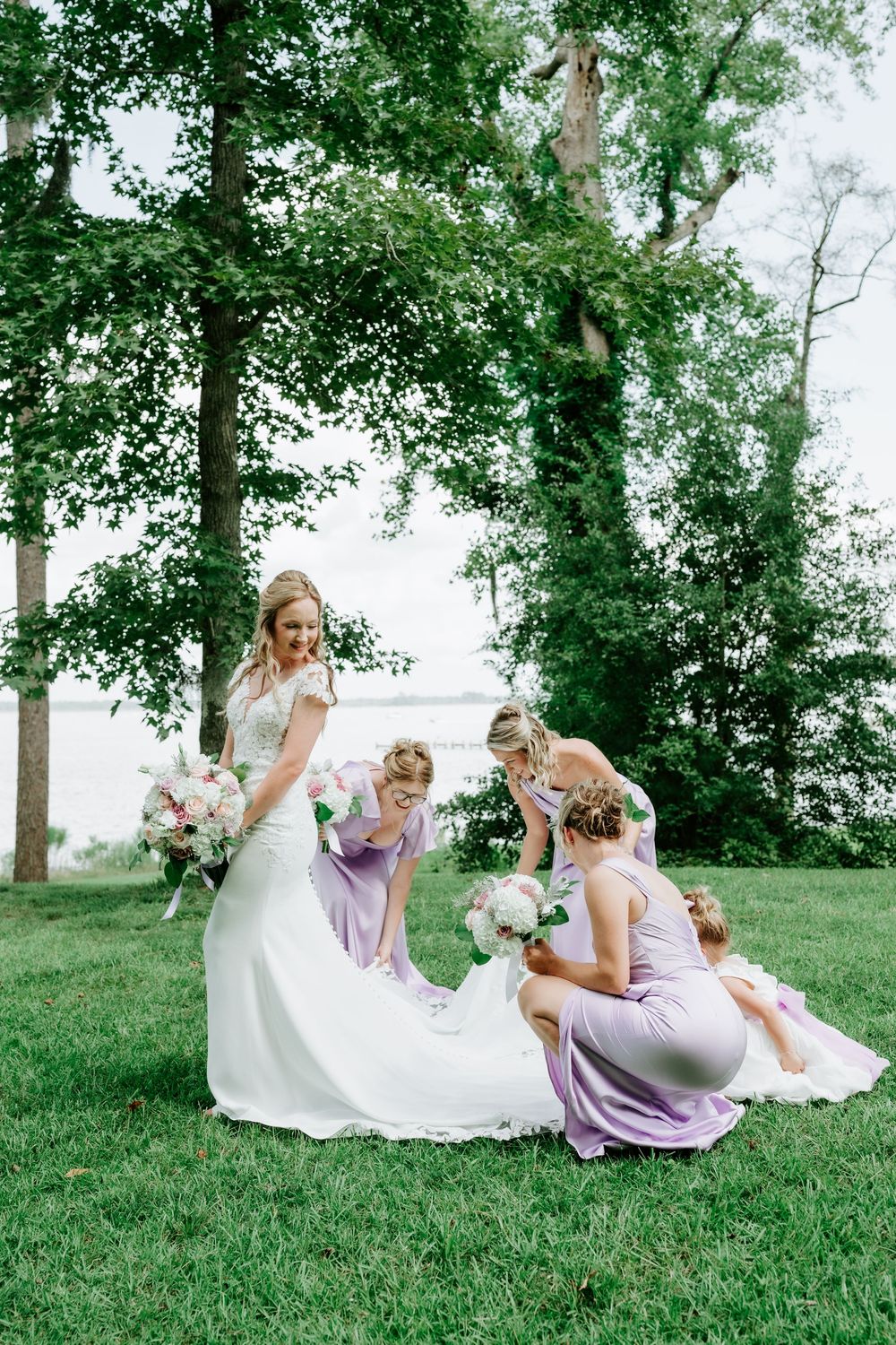 Bride in white gown with bridesmaids in lavender dresses arranging her train on green grass outdoors.