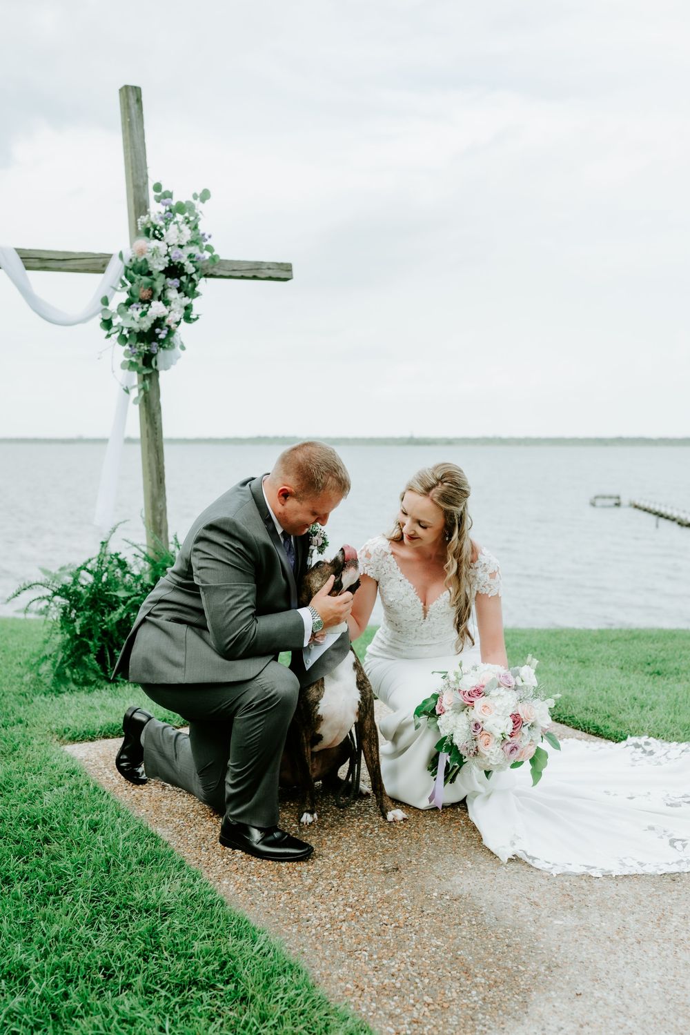 Bride and groom kneel together with their dog near a floral cross altar by the waterfront on their wedding day.