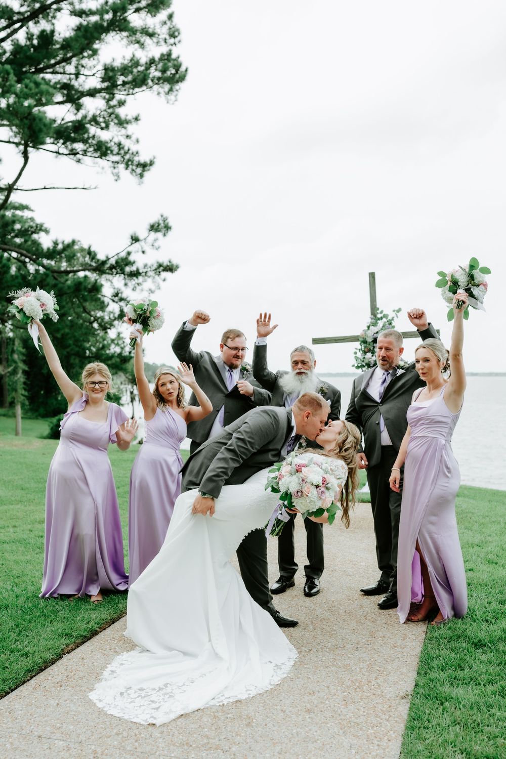 Groom dips bride for a kiss surrounded by cheering wedding party in lavender dresses near a lakeside cross.