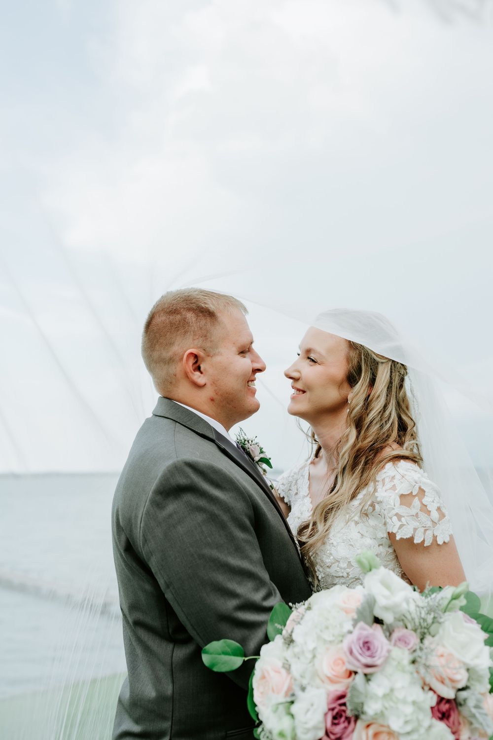 Bride and groom share a romantic moment by the ocean, bride holding a pastel floral bouquet in a lace wedding gown.