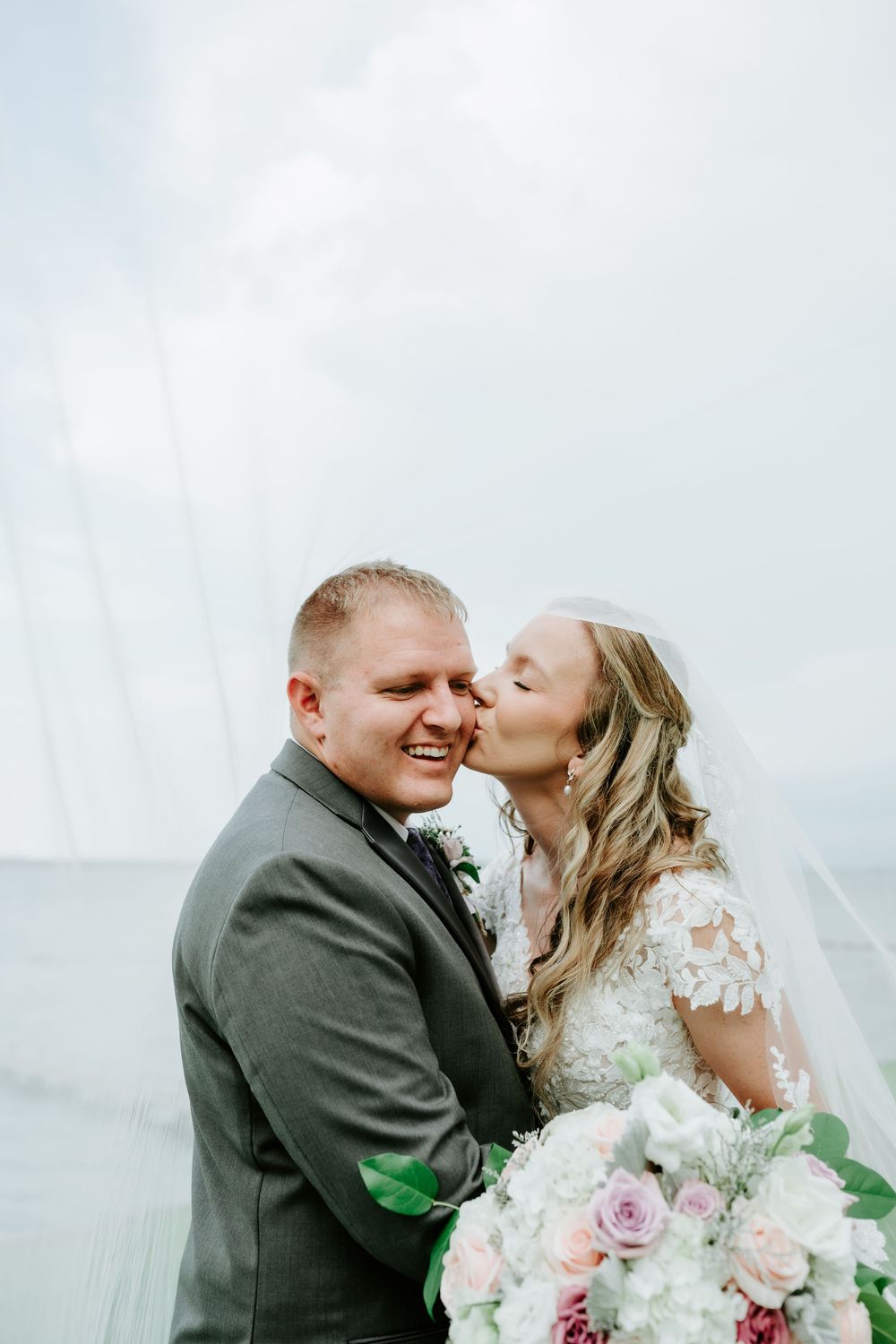 Bride kisses groom on cheek as he smiles, holding a pink and white bouquet with water in background.