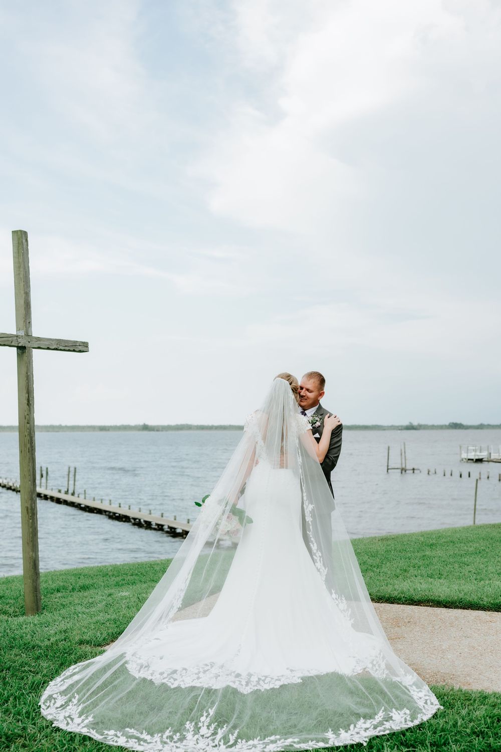 Bride and groom embrace near waterfront with long lace cathedral veil and wooden cross at outdoor wedding.