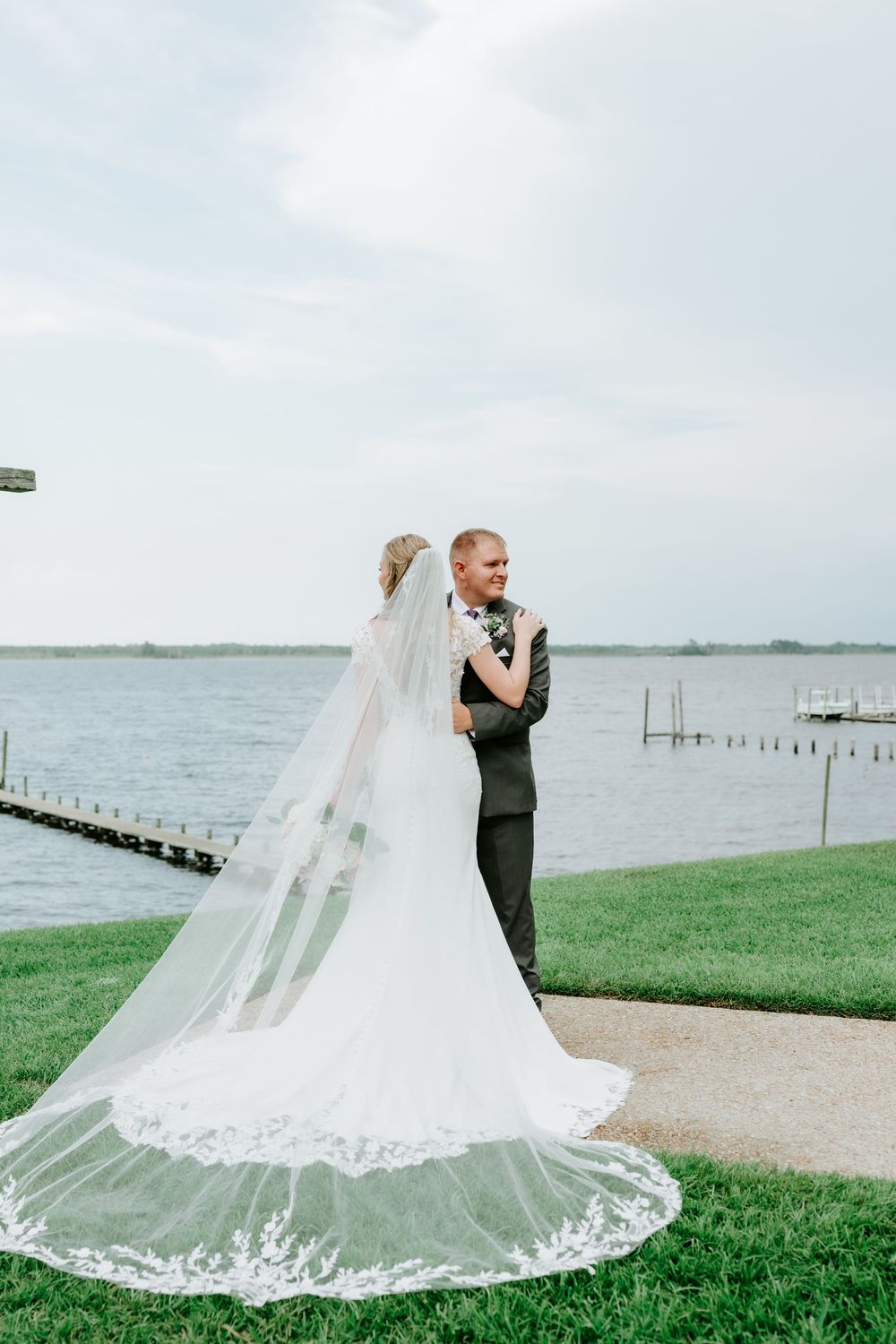 Bride and groom embrace near waterfront, her lace-trimmed cathedral veil trailing on green grass under cloudy skies.