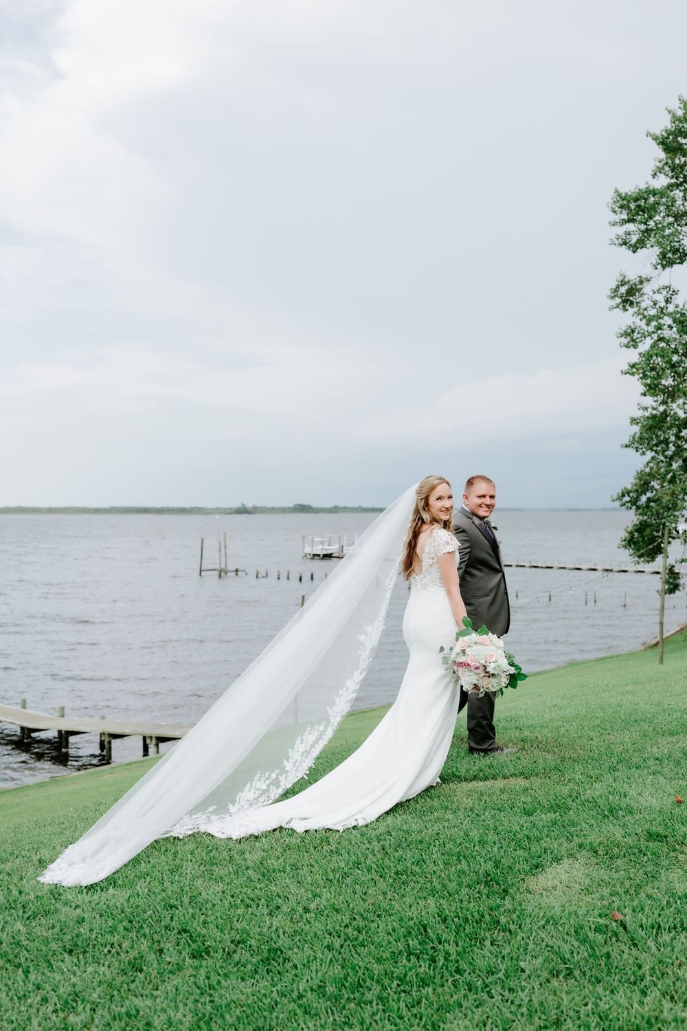 Bride and groom pose by lakeside, her long lace veil flowing in the wind as she holds a bouquet.
