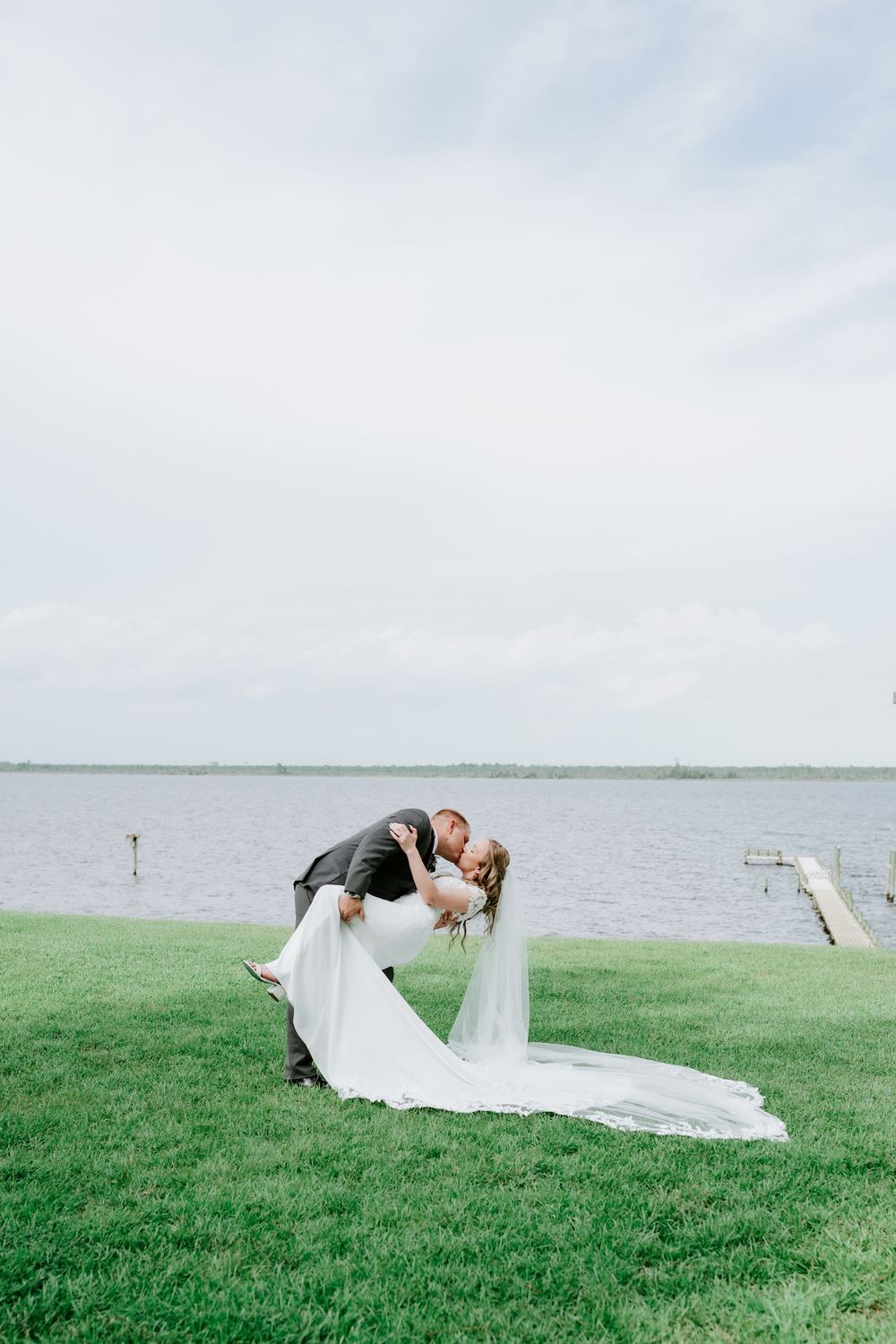 Groom dips bride in a romantic kiss on green lawn by lakeside, her white gown and veil flowing gracefully.