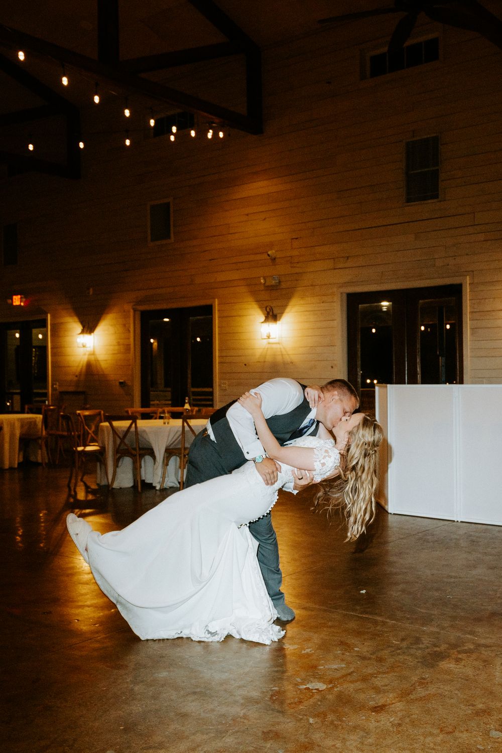 Groom dips bride for a romantic kiss during their first dance in a warmly lit rustic wedding reception hall.