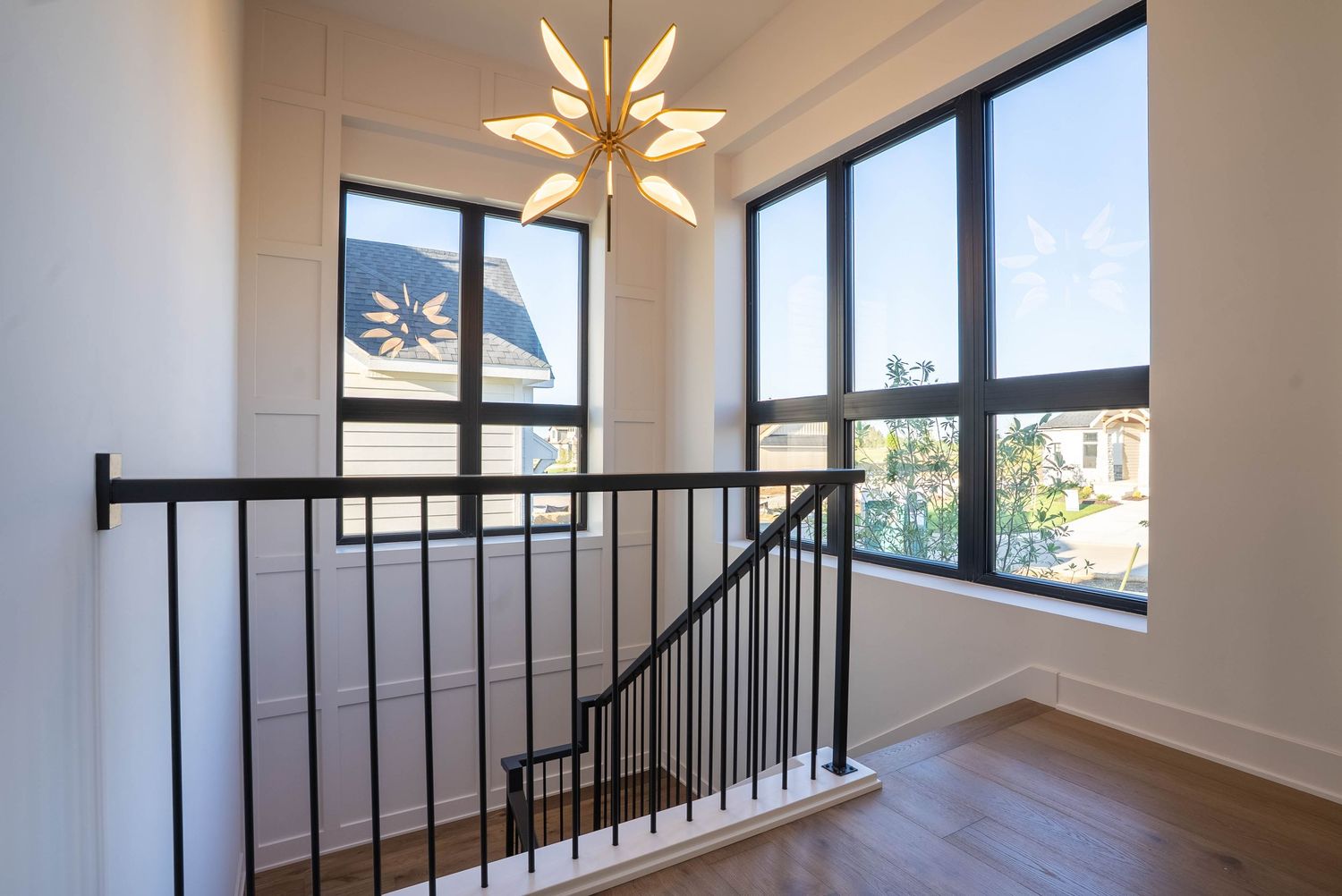 Modern staircase landing with black metal railing, hardwood floors, large windows, and a gold starburst chandelier.