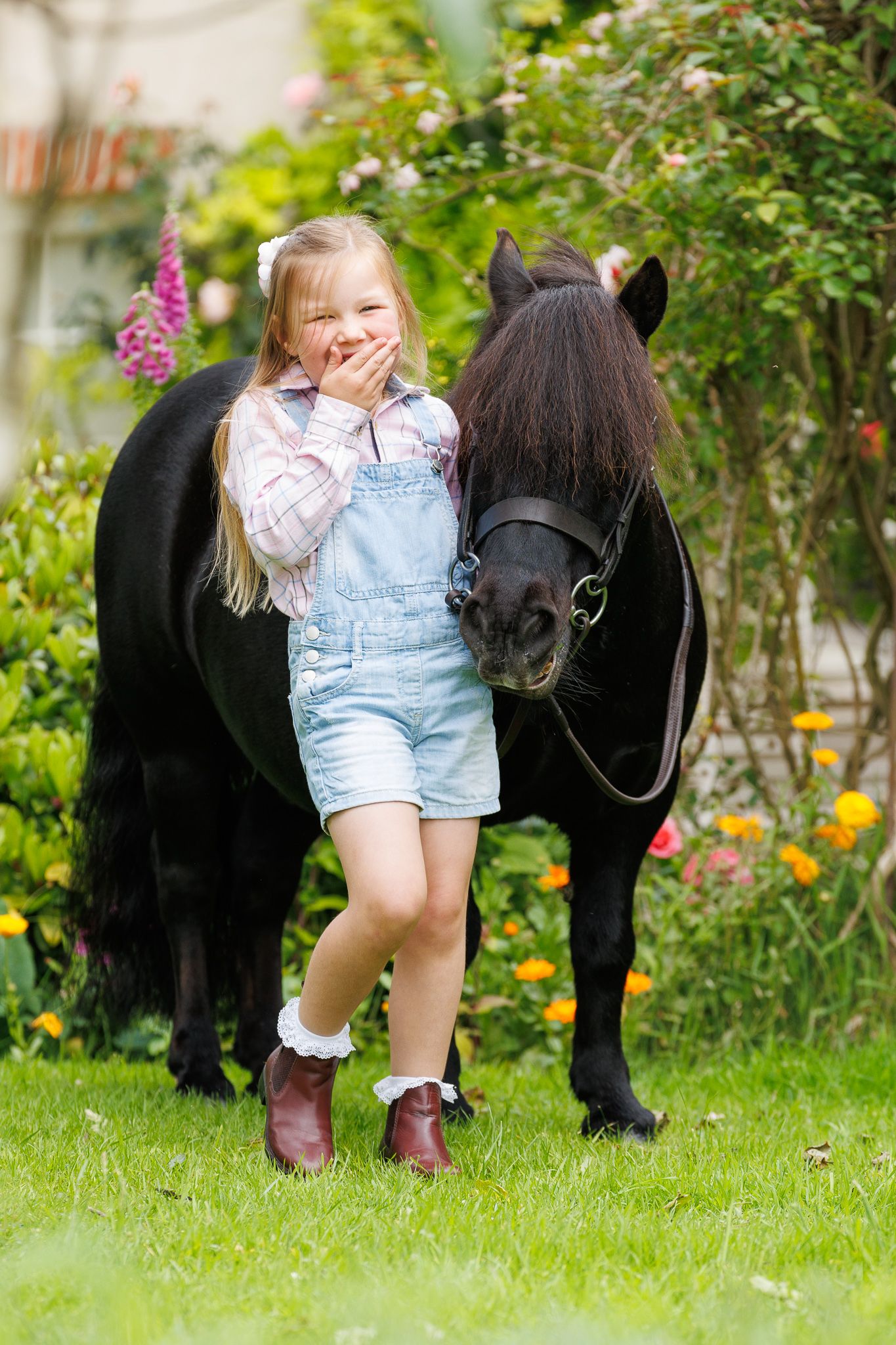 Young blonde girl in white dress hugging a black pony in a lush green garden with daisies.