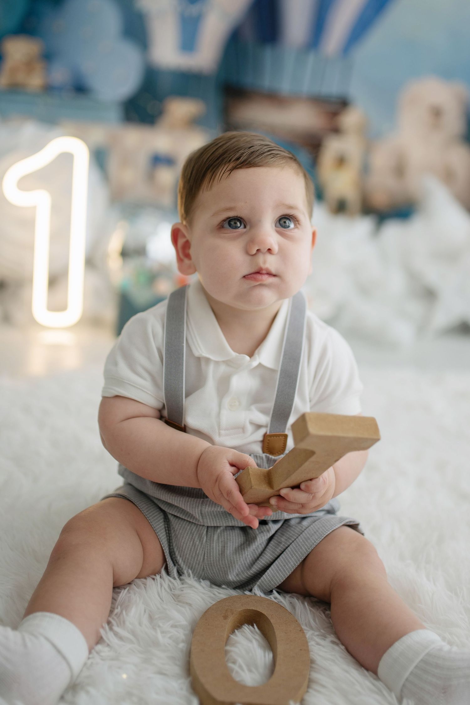 Adorable baby boy in suspenders sits on white fur rug holding wooden number toys beside glowing number 1.