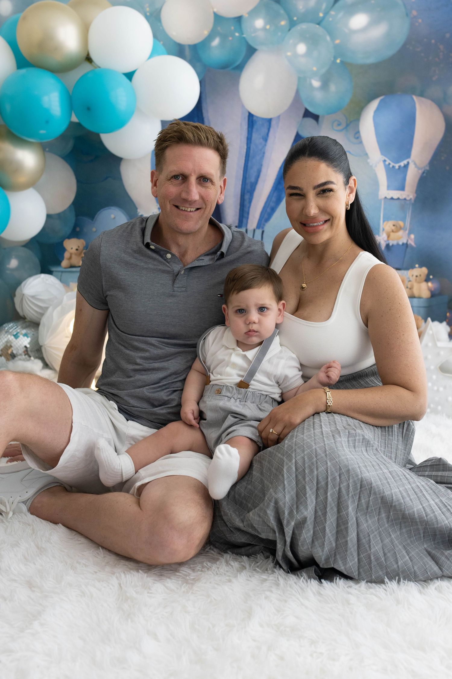 Family photo with parents and baby boy sitting on white fur rug with blue balloon arch backdrop.