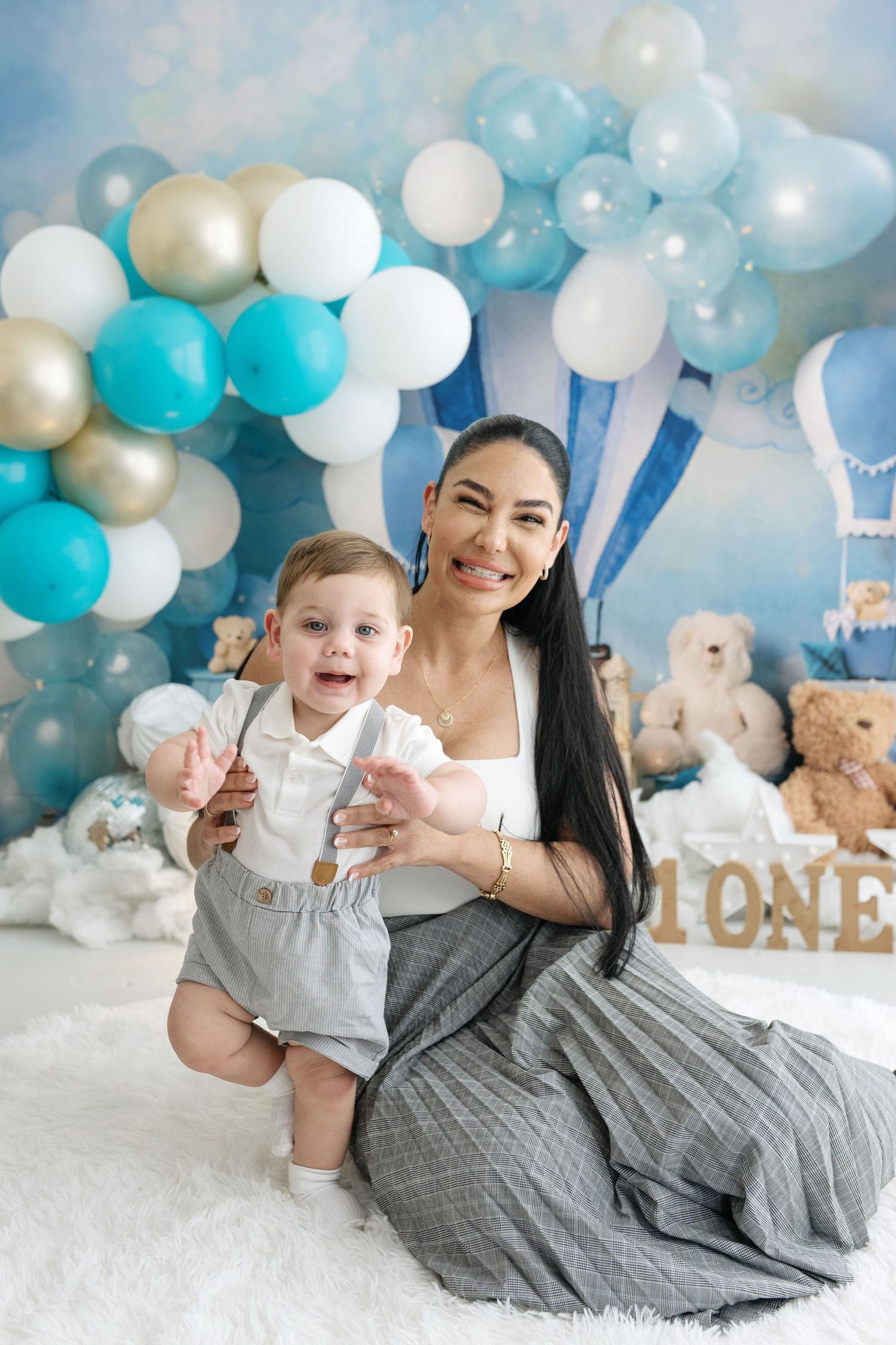 Mother and baby boy celebrate first birthday with blue and gold balloon arch and teddy bears.