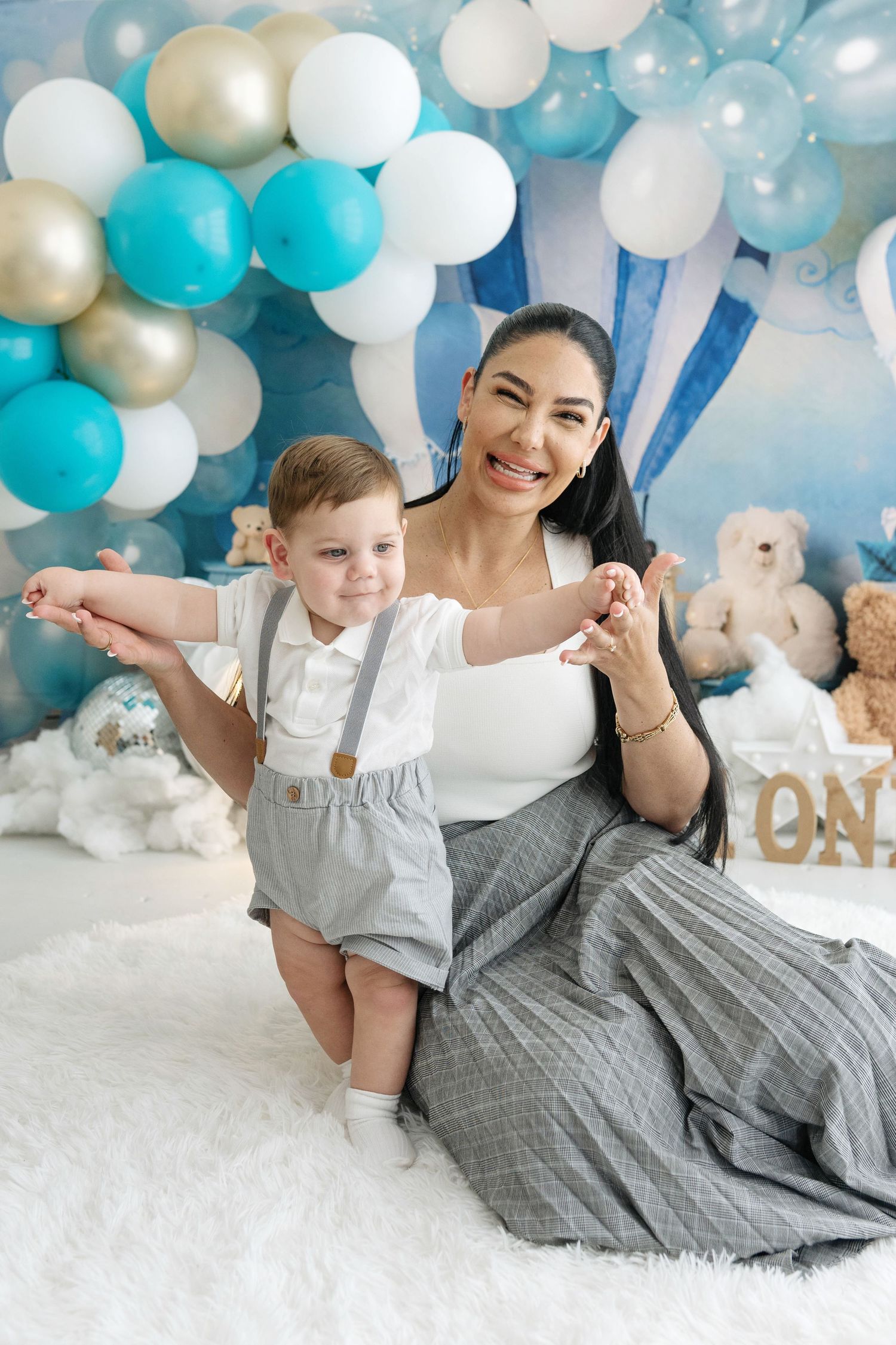 A smiling mother sits with her toddler son at a blue and white balloon-decorated first birthday party photo shoot.
