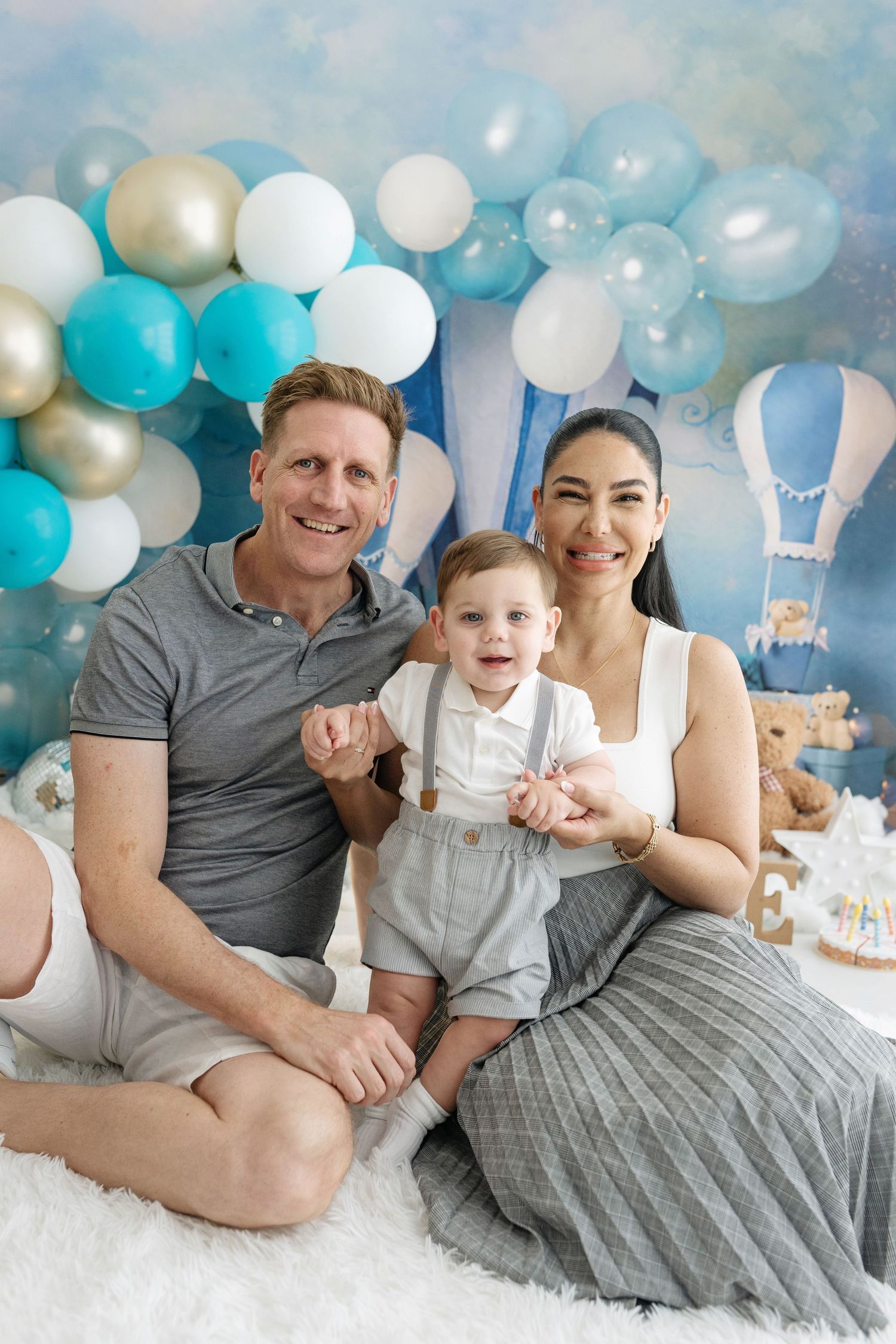 Family portrait with parents and baby boy at first birthday party with blue and white balloon backdrop.