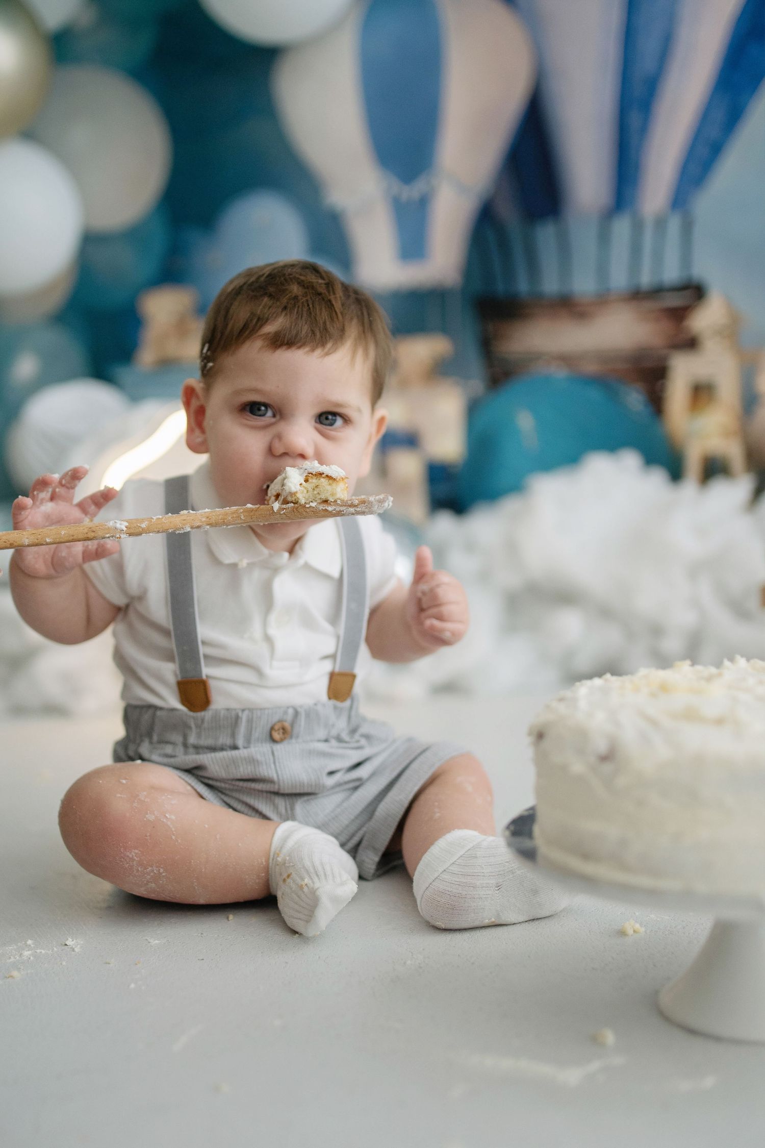 Baby boy in suspenders eating cake at first birthday smash cake party with blue balloon decorations.
