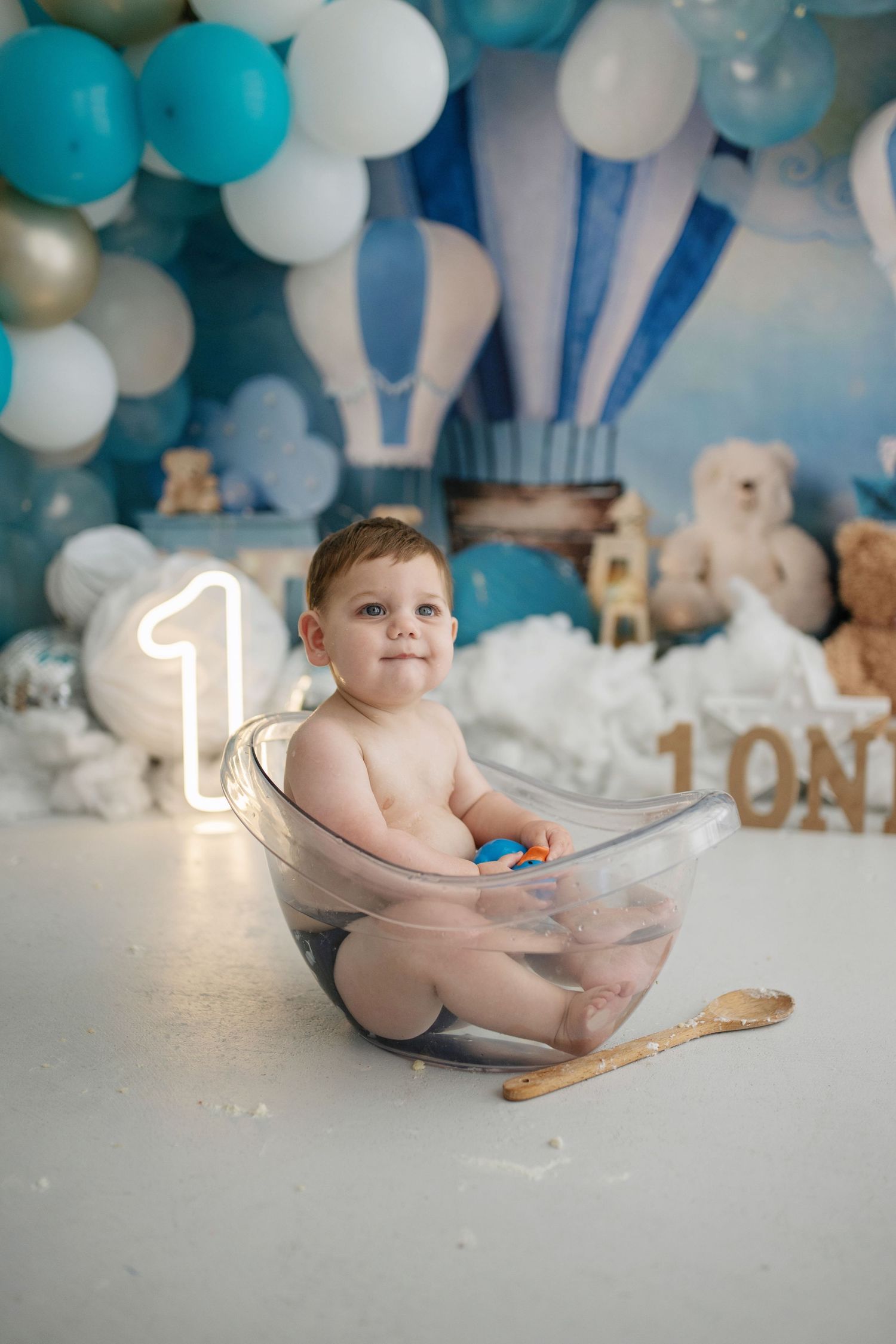 Baby boy sits in clear bowl at 1st birthday cake smash with blue balloons and hot air balloon backdrop.