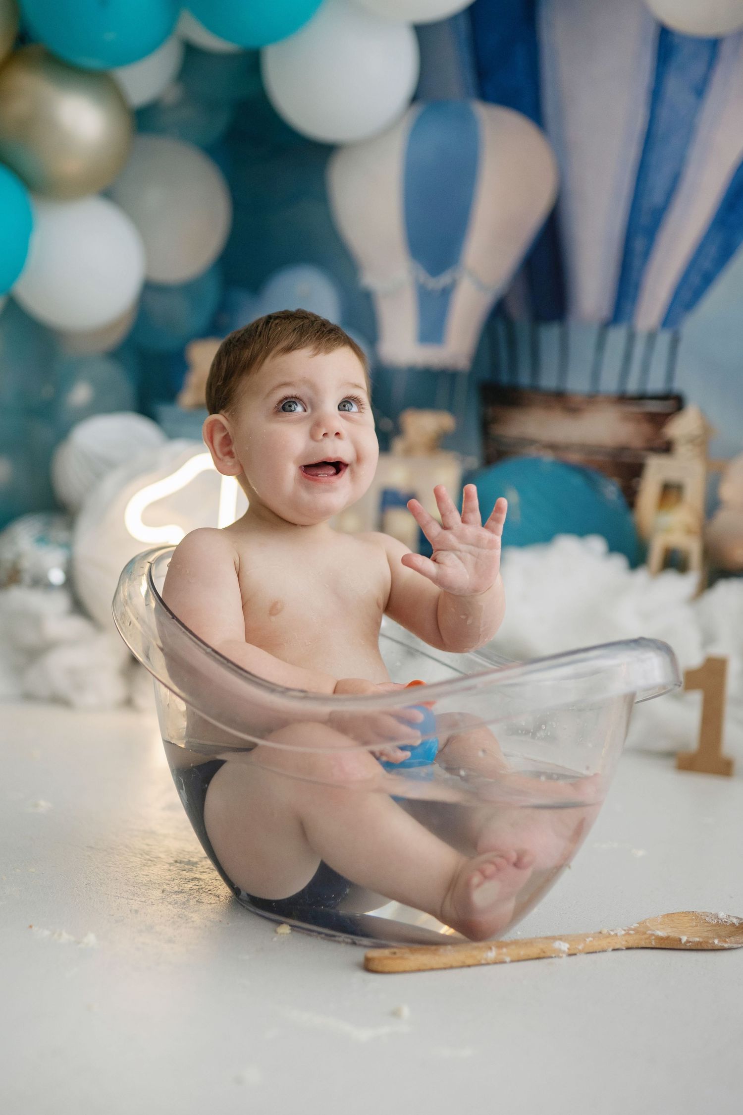 Happy baby boy sitting in a clear tub at a first birthday party with blue balloon decorations.