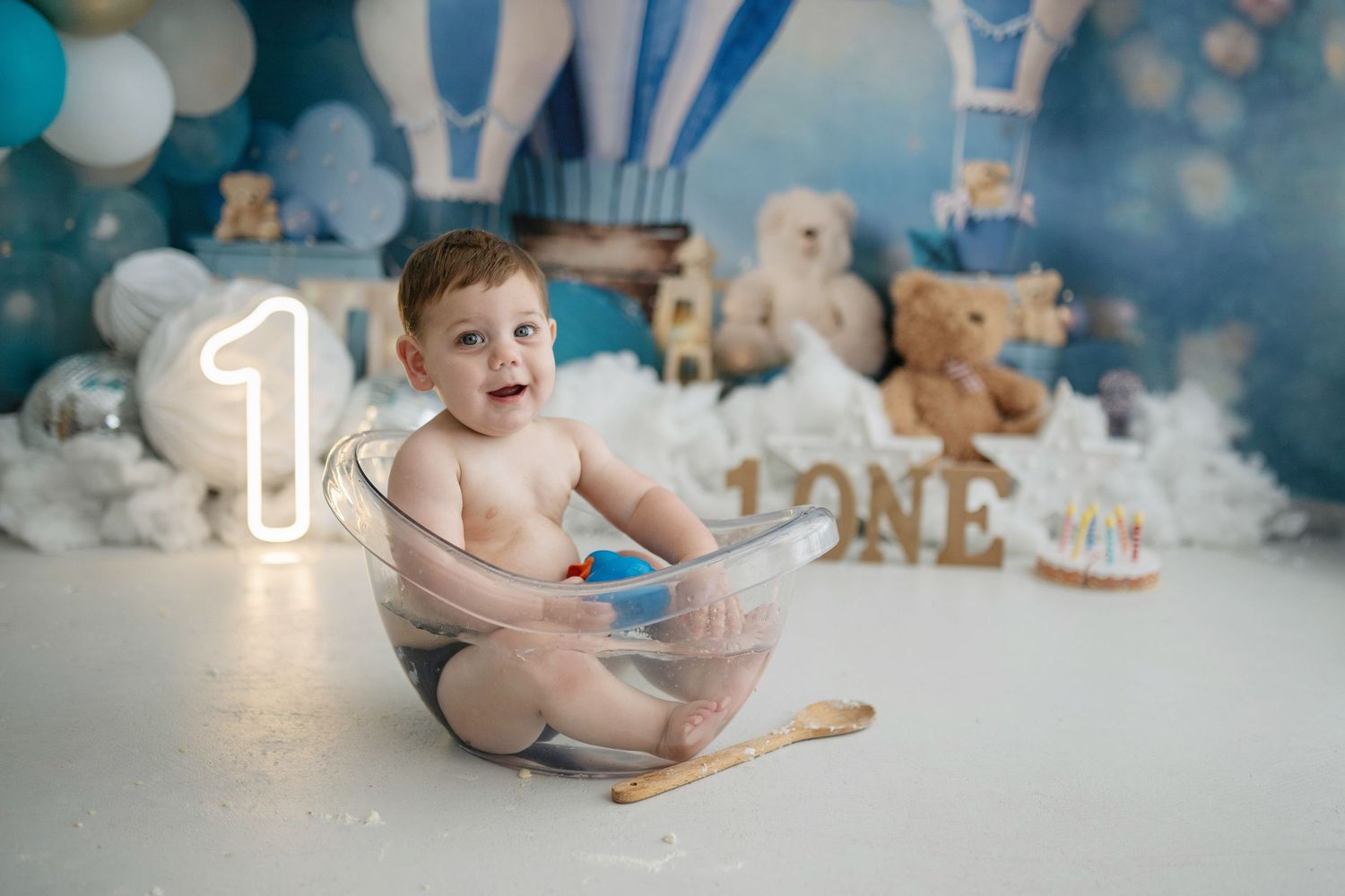 Baby sitting in a clear tub at a first birthday cake smash with blue balloons and teddy bears.