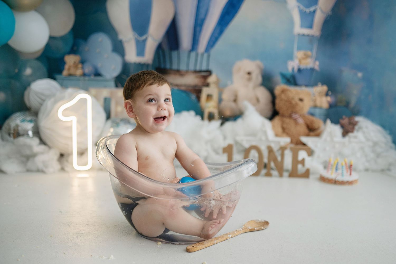 Baby boy sitting in a clear tub during a first birthday cake smash photoshoot with blue balloon decorations.