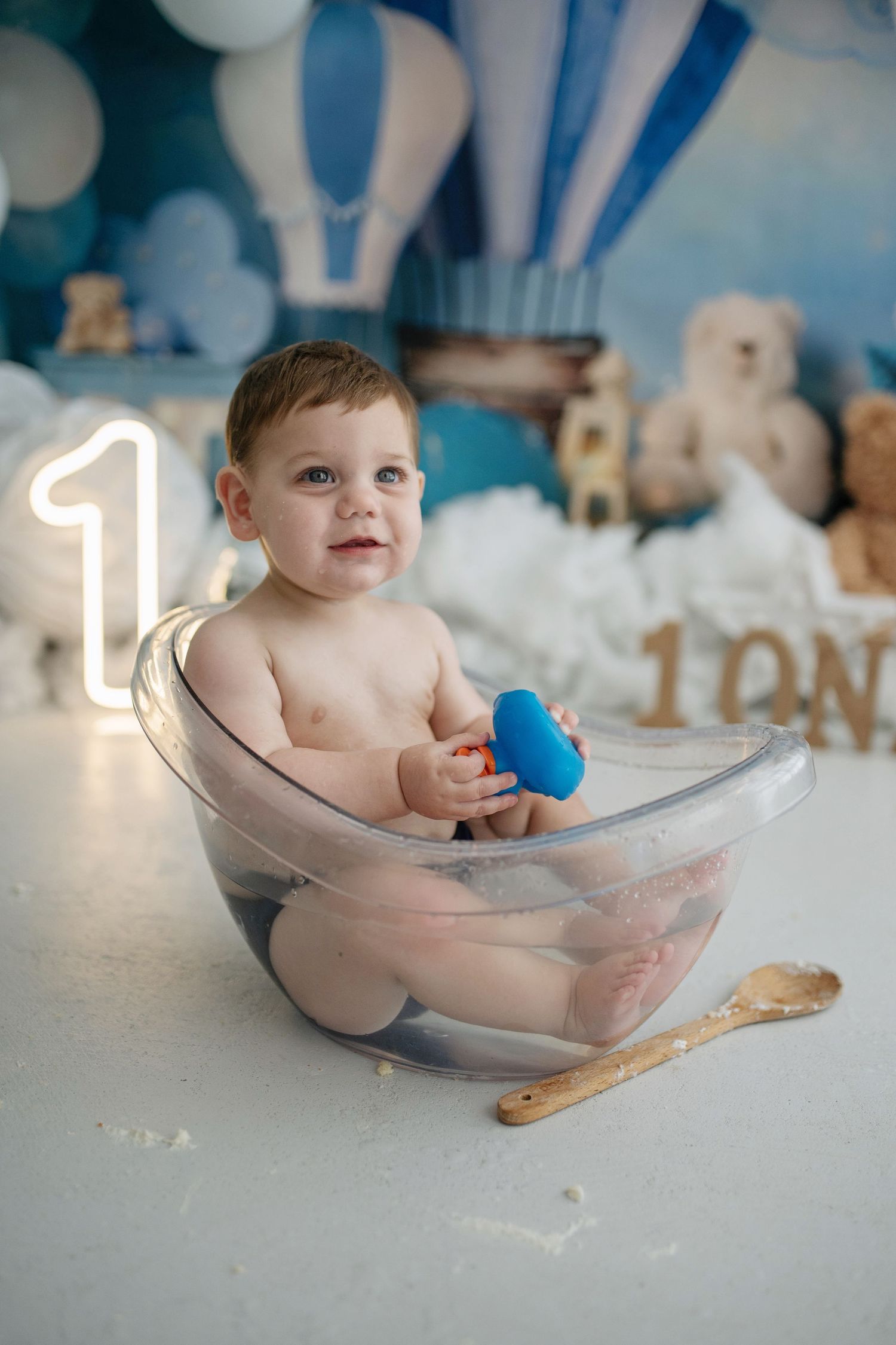 Baby boy sits in clear tub holding blue toy at first birthday cake smash with hot air balloon decor.