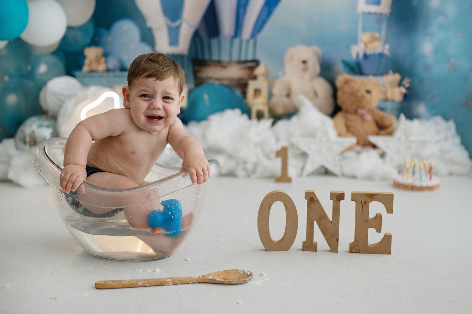 Baby's first birthday cake smash photo with teddy bears, balloons, and ONE sign, sitting in glass bowl.