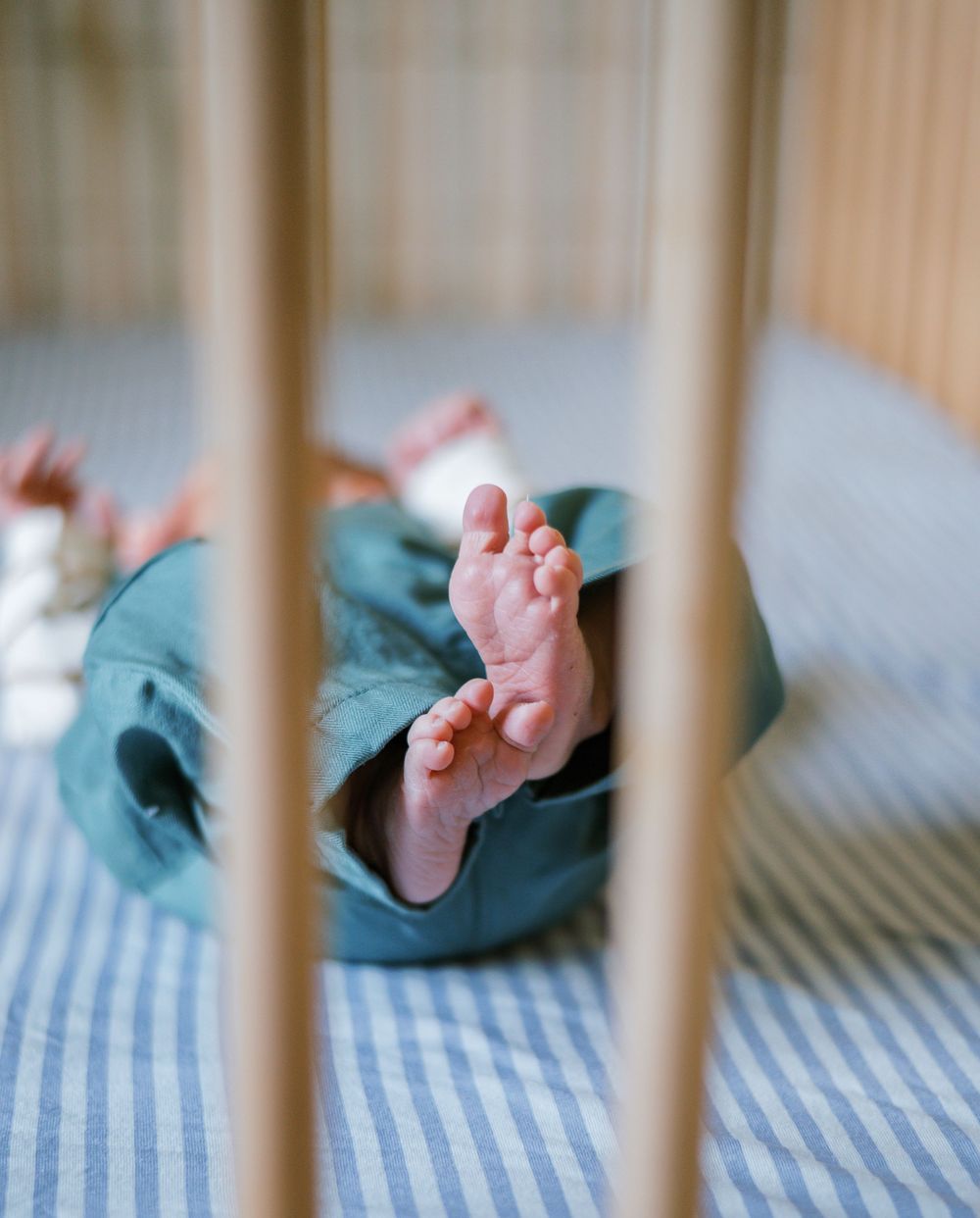 Newborn baby tiny feet peeking through wooden crib slats on a striped blue mattress.