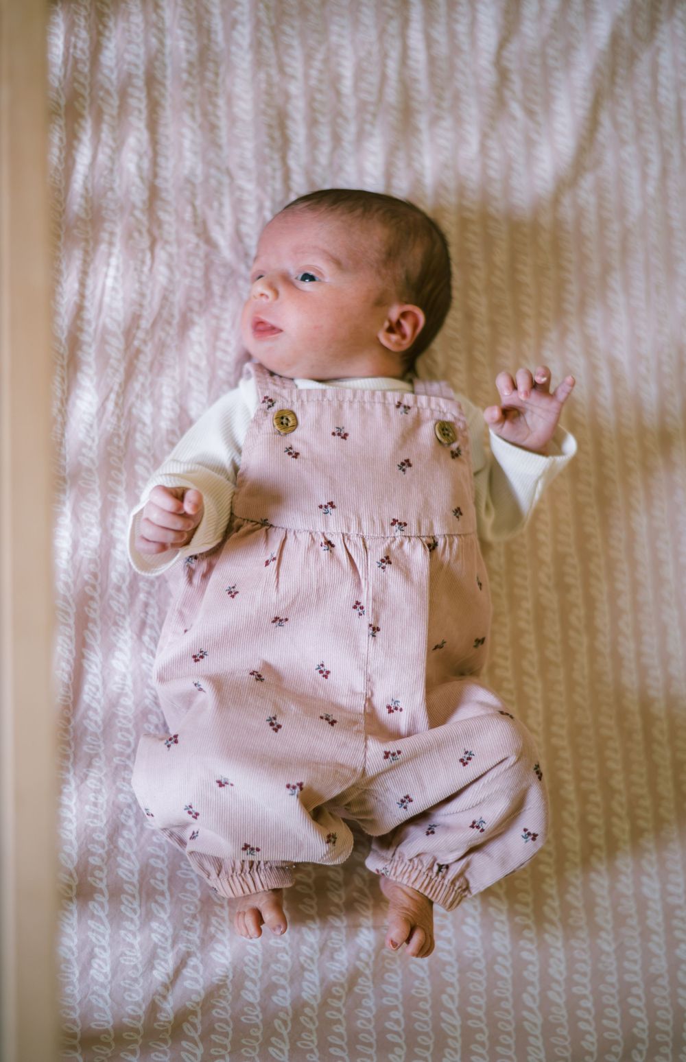 Newborn baby in pink floral dungarees lying on a white textured blanket indoors.