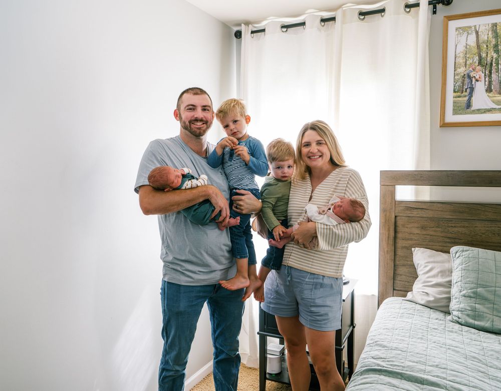 Happy family of four posing indoors, parents holding newborn twins and toddler, smiling near a bed in a bright bedroom.