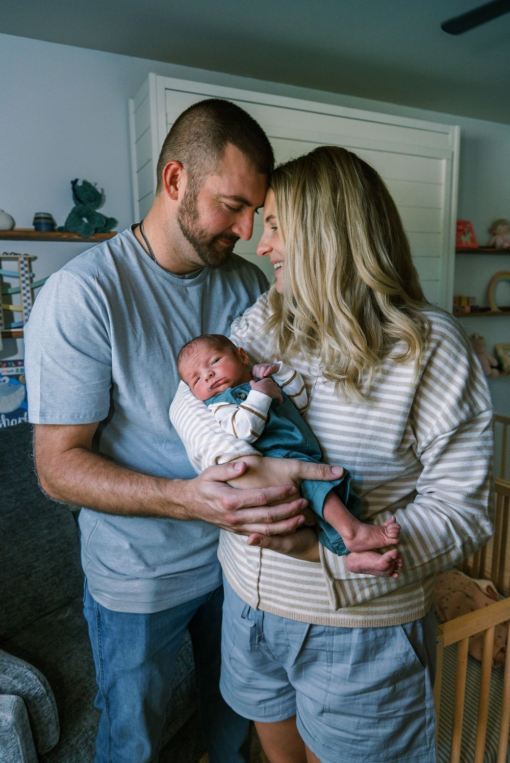 New parents share a tender moment, touching foreheads while cradling their newborn baby in a cozy nursery room.