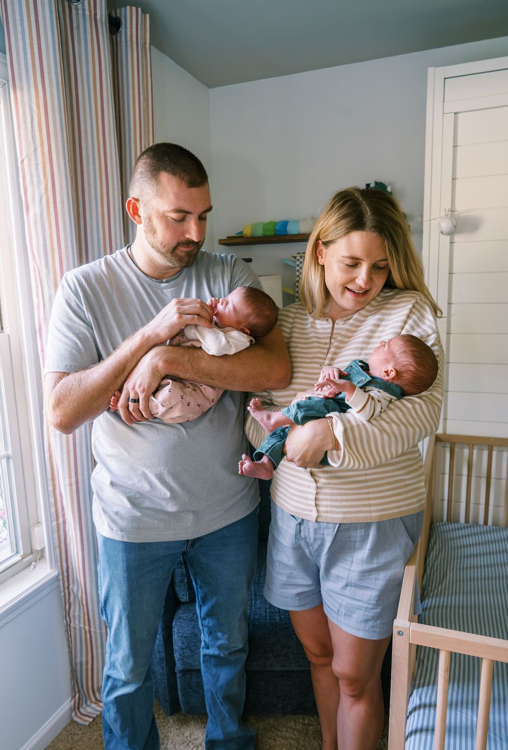 New parents holding newborn twins in a nursery room, smiling lovingly at their babies.
