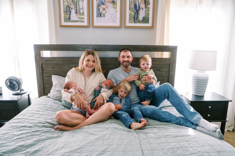 Happy family with young children posing together on a bed in a bright bedroom with framed photos on the wall.