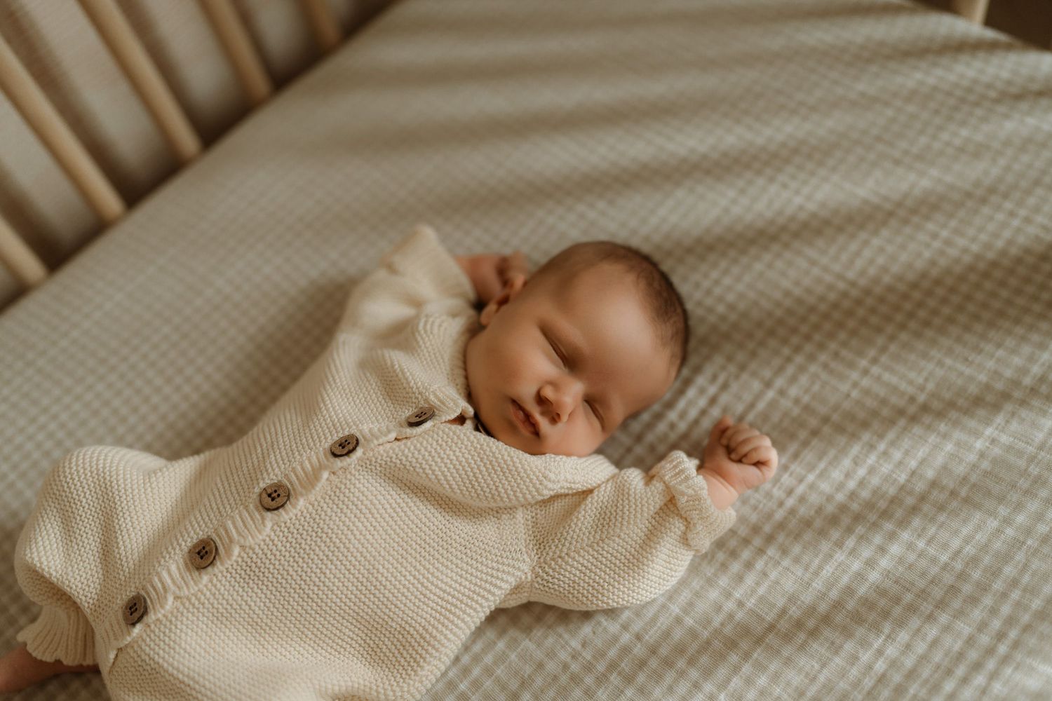 Newborn baby sleeping peacefully in a crib wearing a cream knit cardigan on a soft checkered sheet.