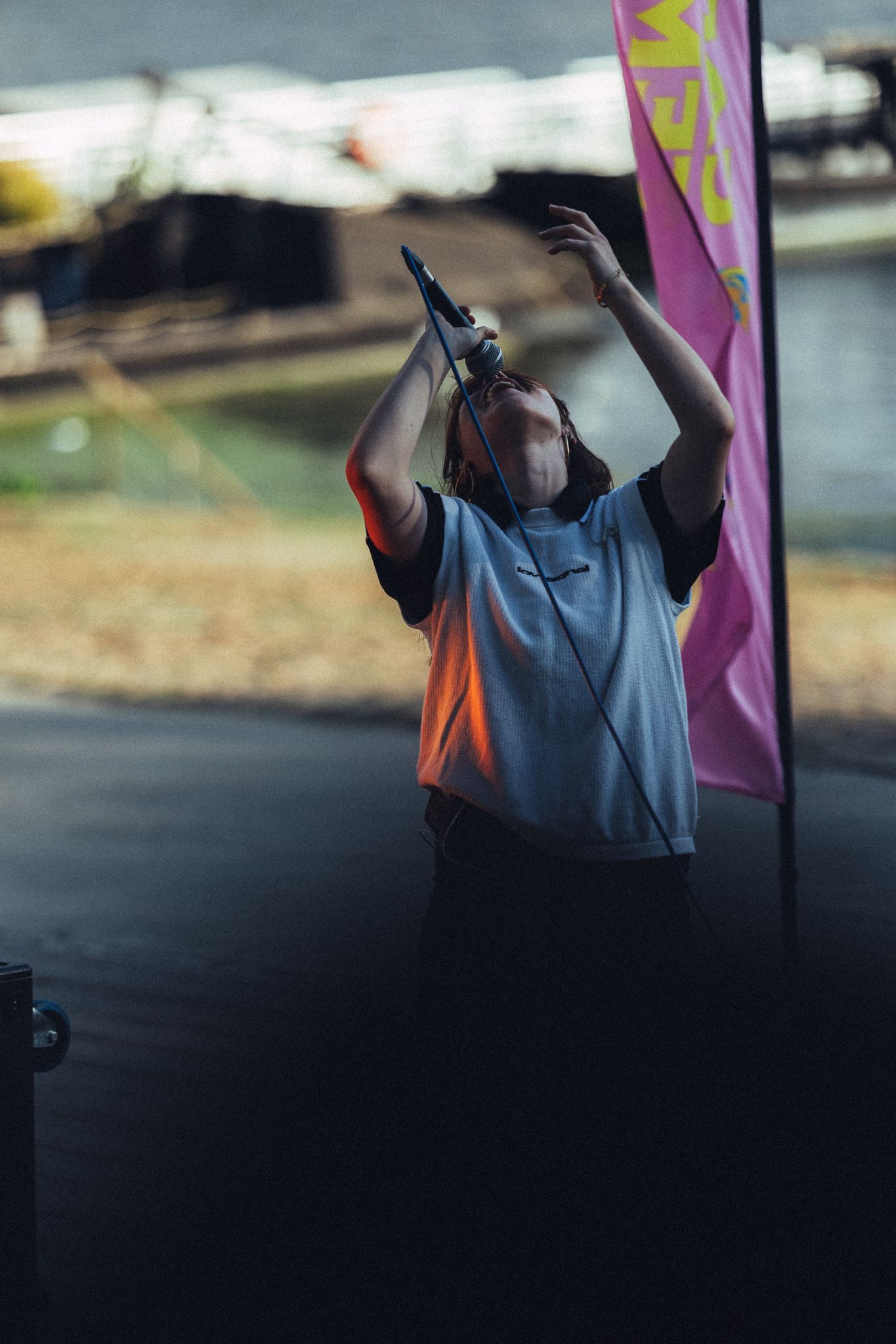 Performer singing on outdoor stage near waterway, arms raised holding microphone, beside pink promotional flag.