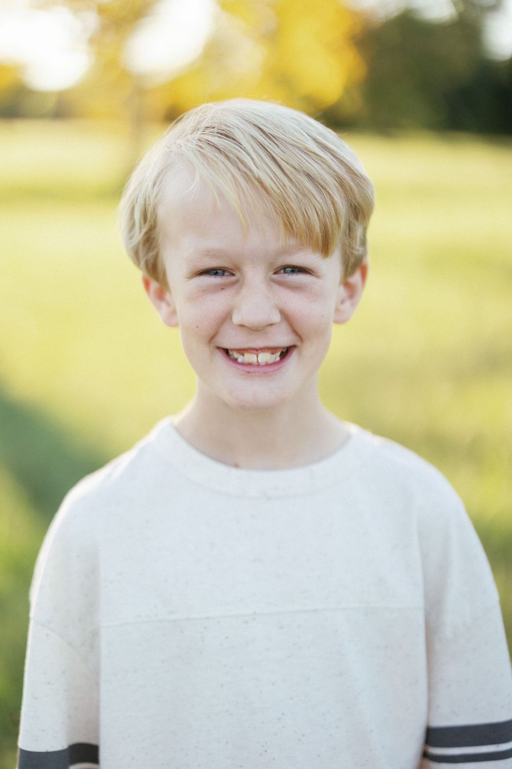 Smiling blond boy in white striped t-shirt posing outdoors in a sunny green field.