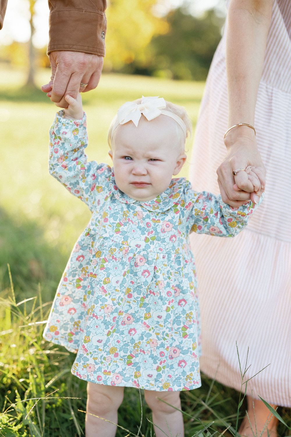 Adorable baby girl in floral dress holding parents hands while standing in a sunny park.
