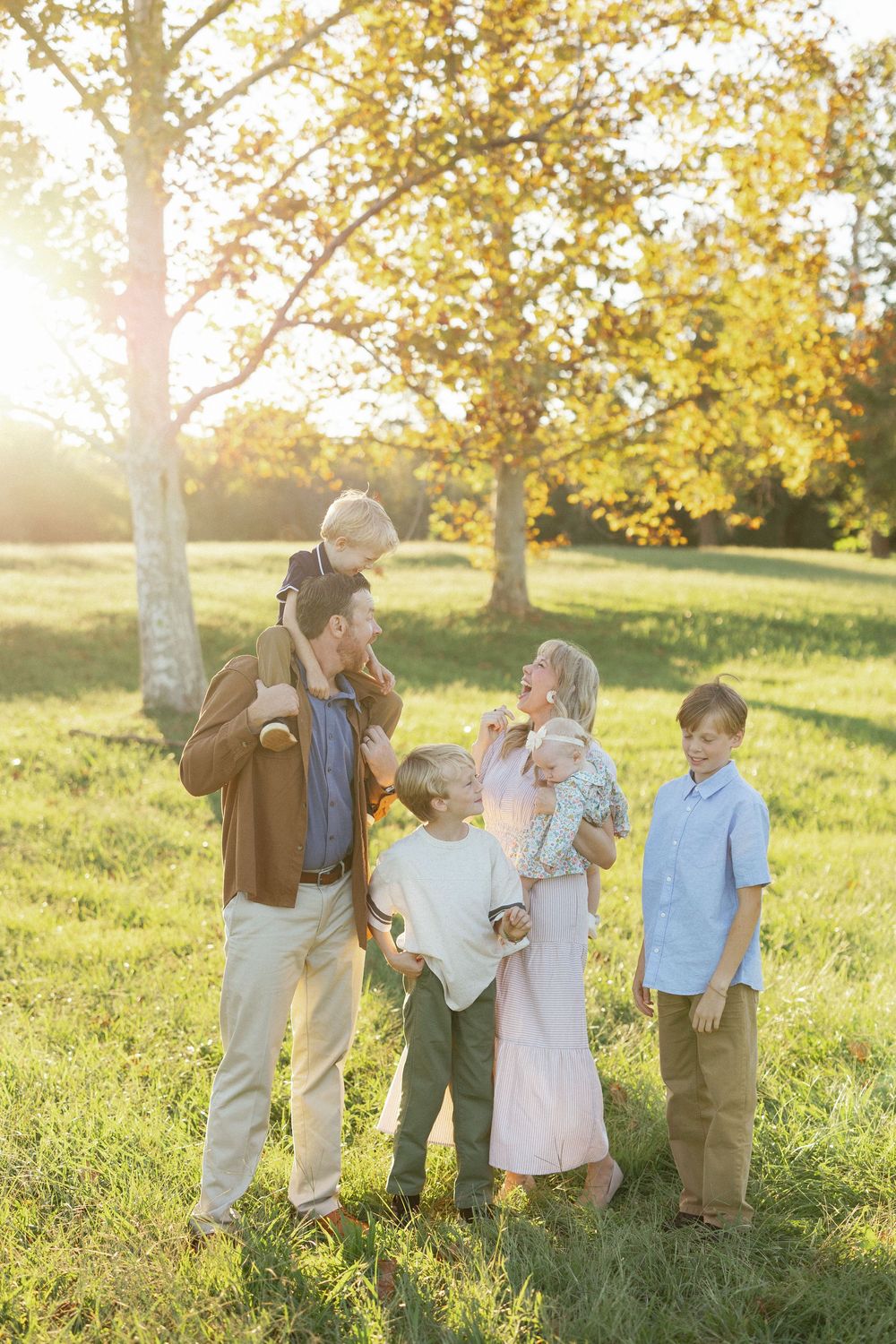 Family of six enjoying a sunny autumn day outdoors, with parents holding children among golden trees.